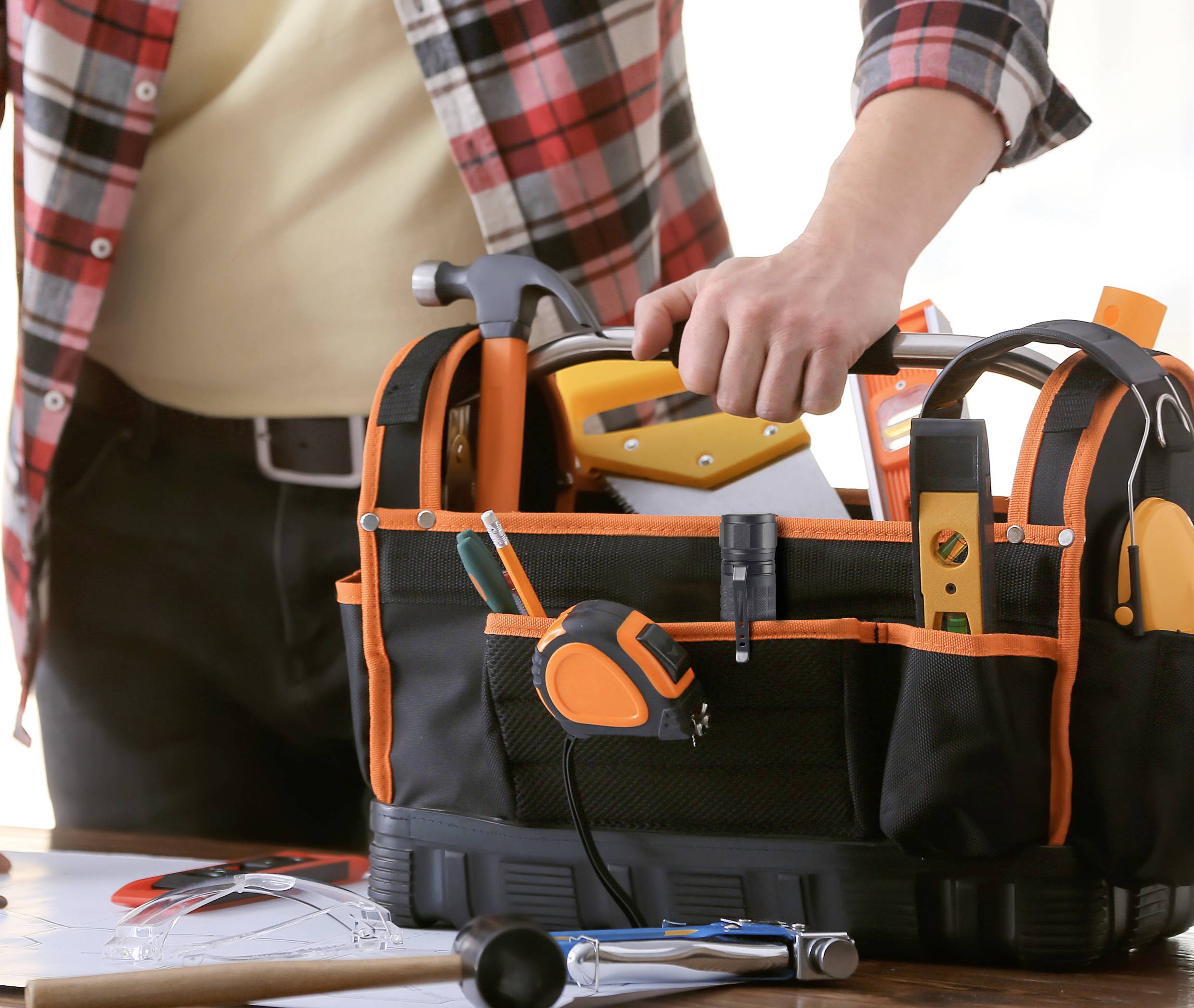 A person is packing tools into a black tool bag with orange details. Plans and a hammer are lying on the table.