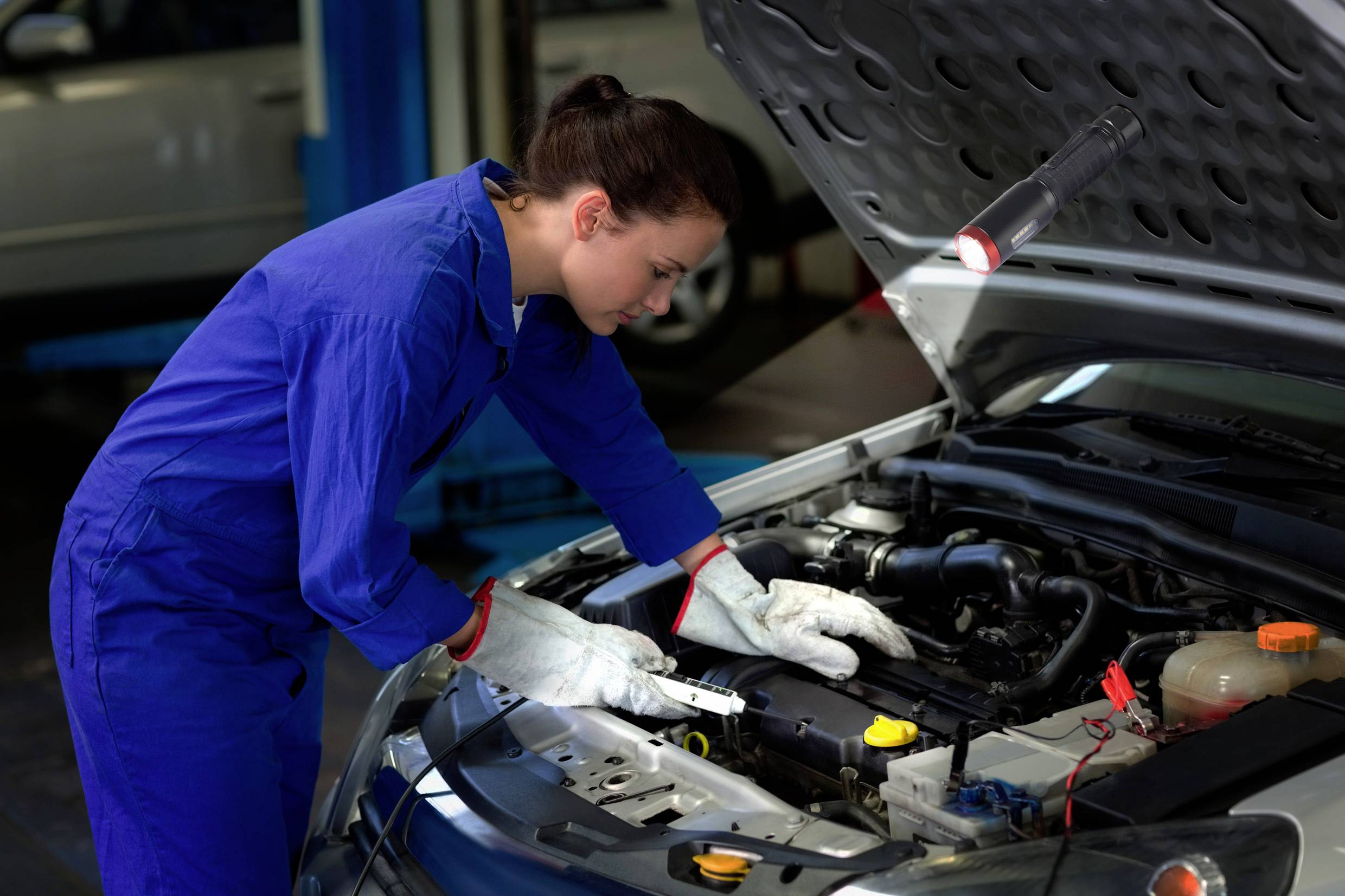 A person in a blue overall is examining the engine of a car in a workshop.