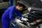 A person in a blue overall is examining the engine of a car in a workshop.