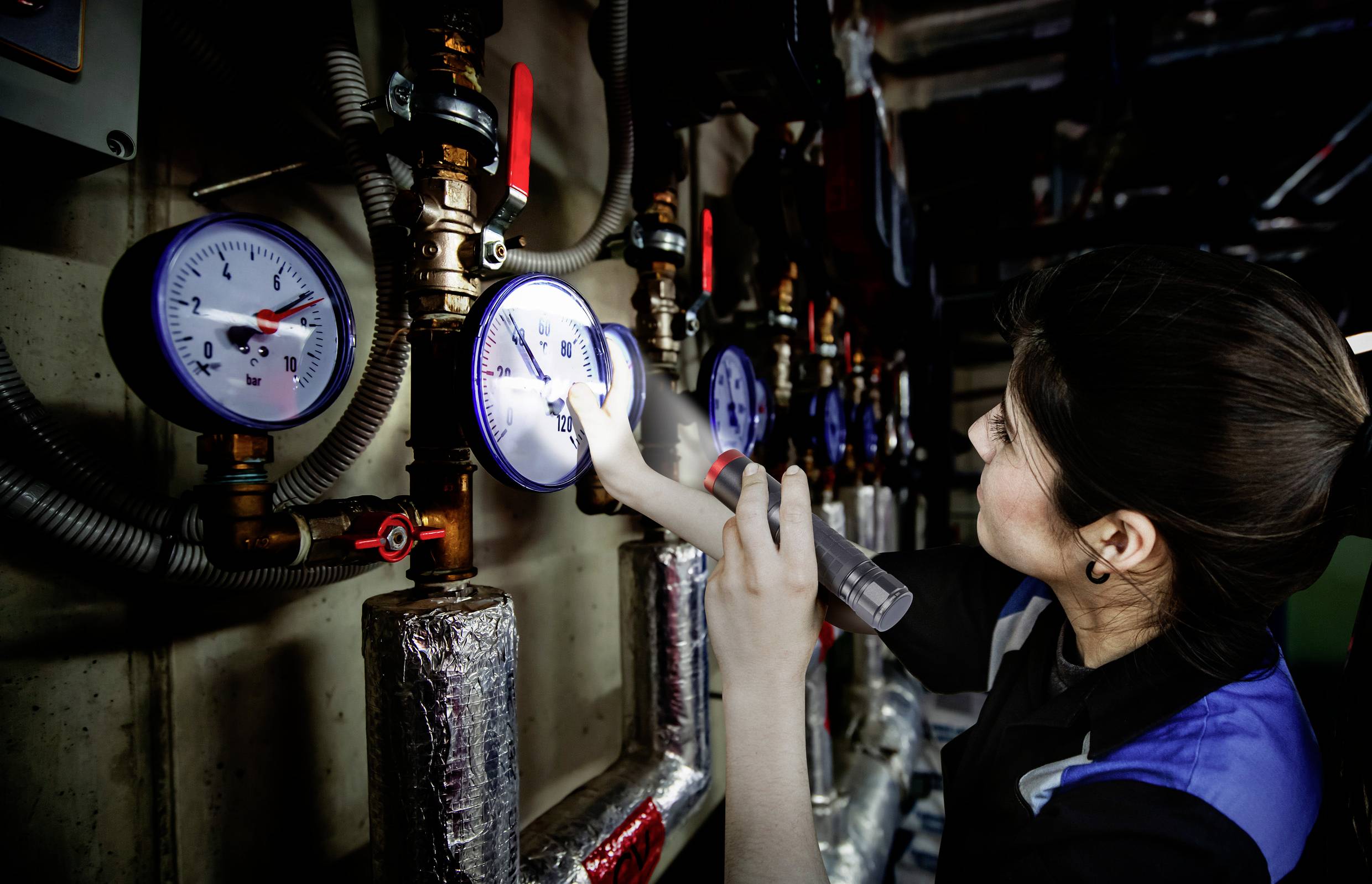 A person is checking several pressure indicators on industrial pipelines and valves using a torch.