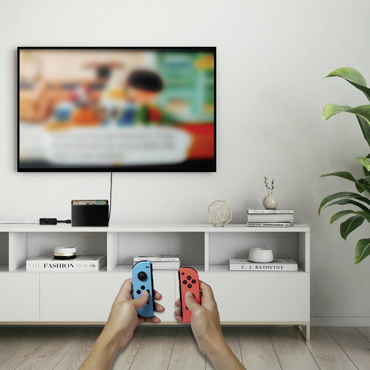 A television displays a video game. Two hands are holding colourful controllers. A white shelf with decorations stands in the background.