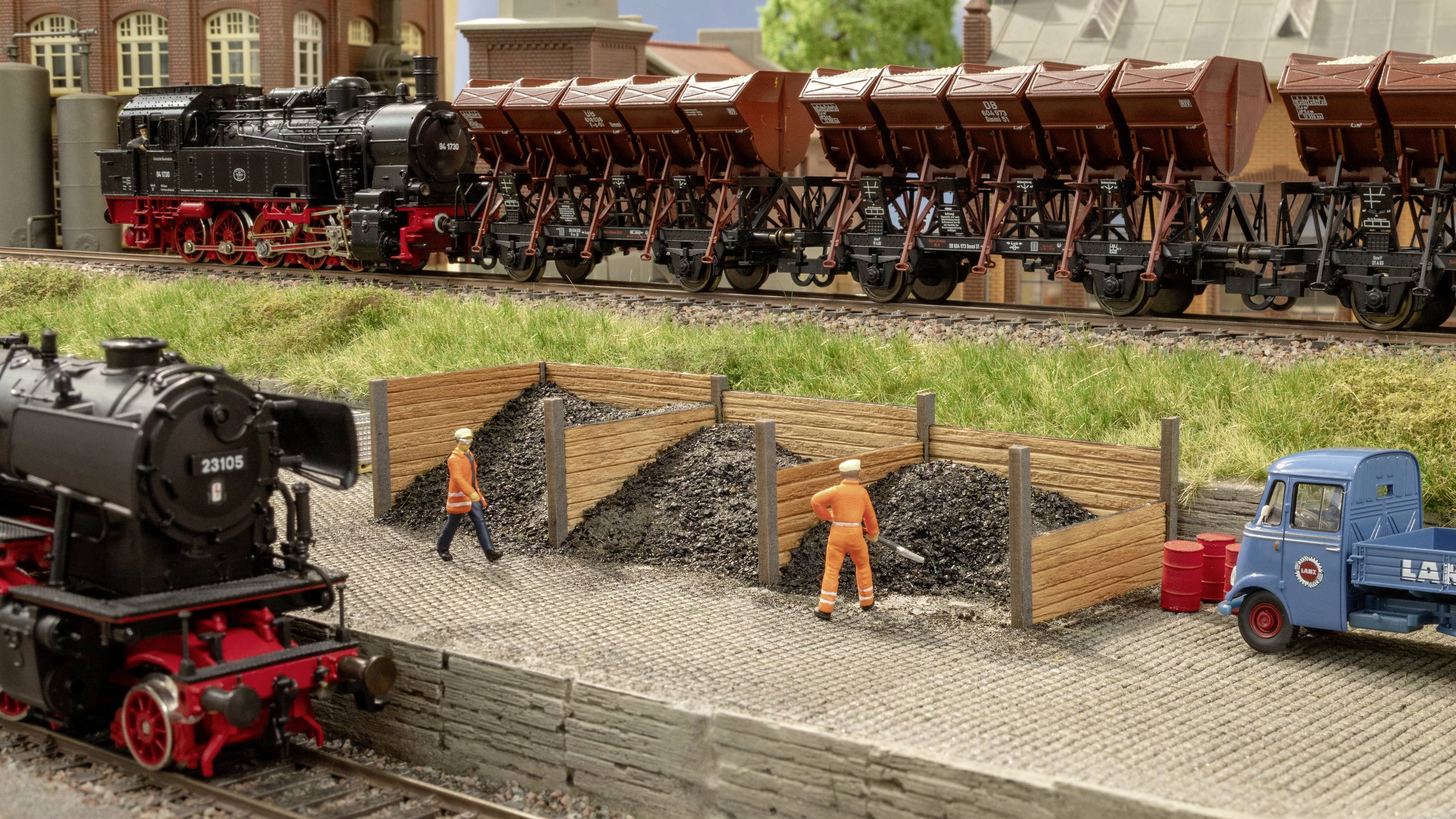 Model railway scene with two steam locomotives and a blue delivery van, two figures in workwear moving coal on the platform.