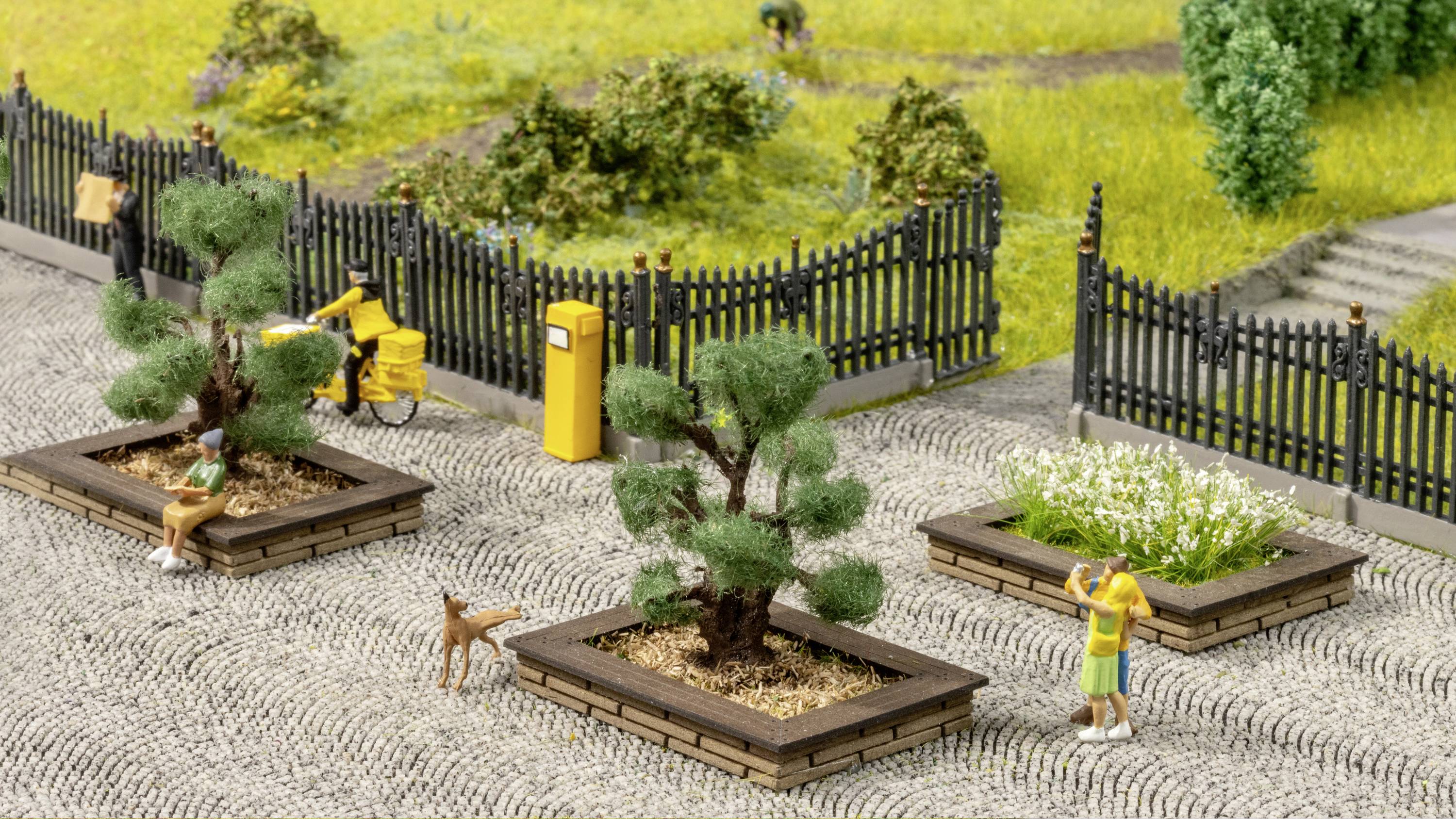 A miniature park with two people on a paved area. One figure is standing next to a tree, the other is walking a dog on a lead. In the background, a fence and flowering plants can be seen.