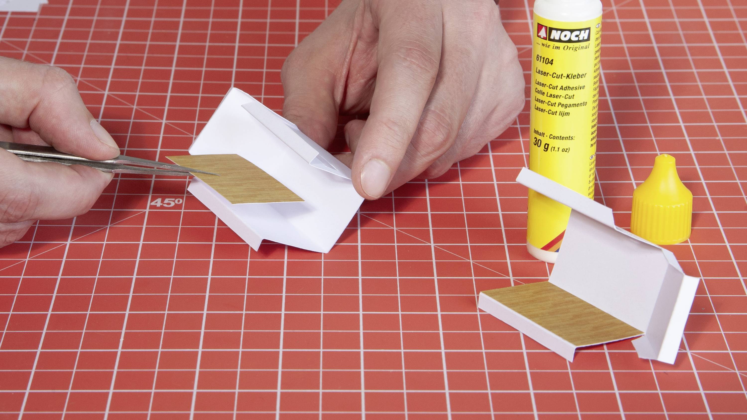 Two hands are constructing a miniature floor on a red and white chequered surface, using glue and tweezers.
