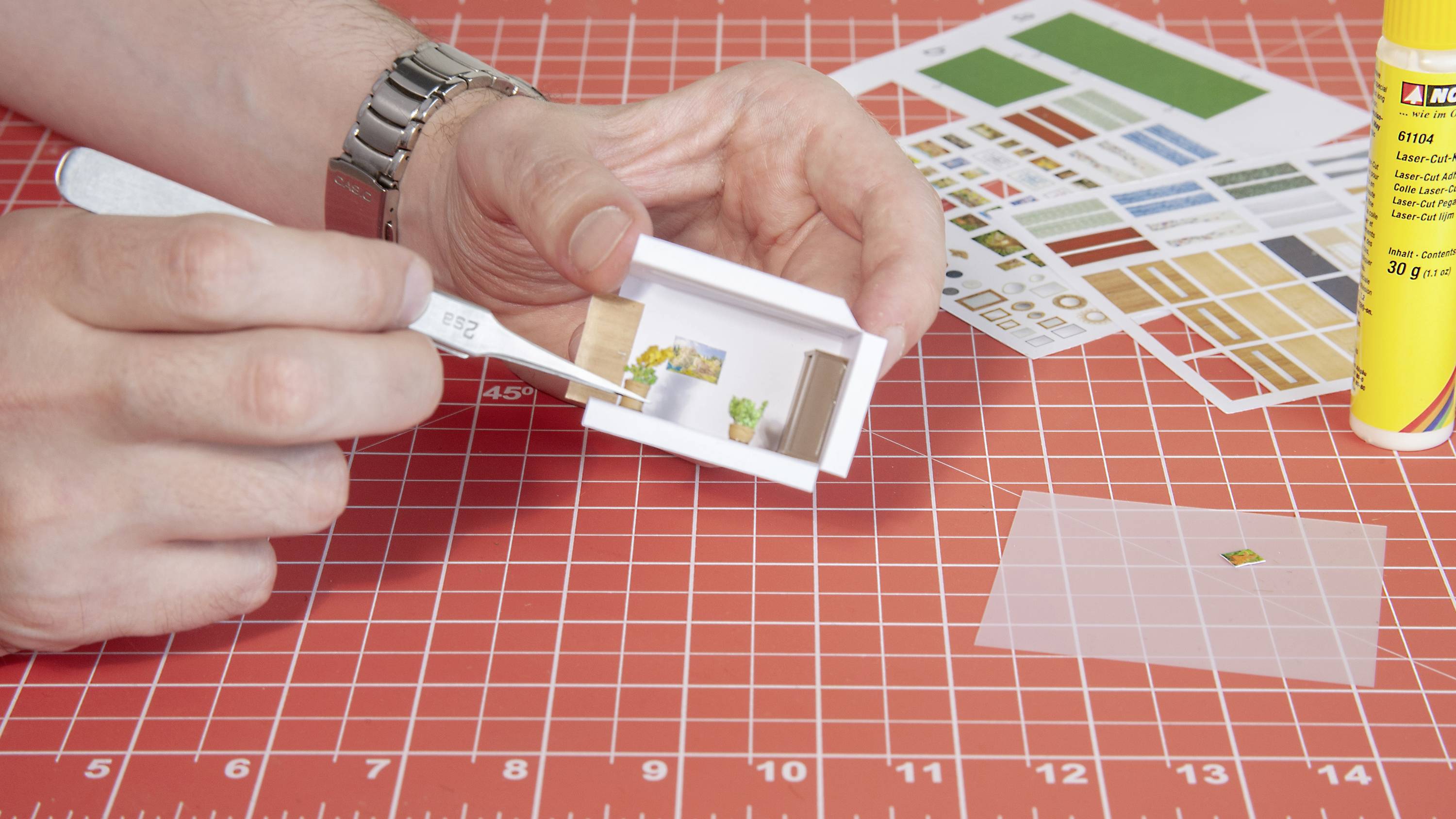 A person is crafting a miniature model of a living space using paper ingredients on a red cutting board. A tube of glue is positioned to the right.
