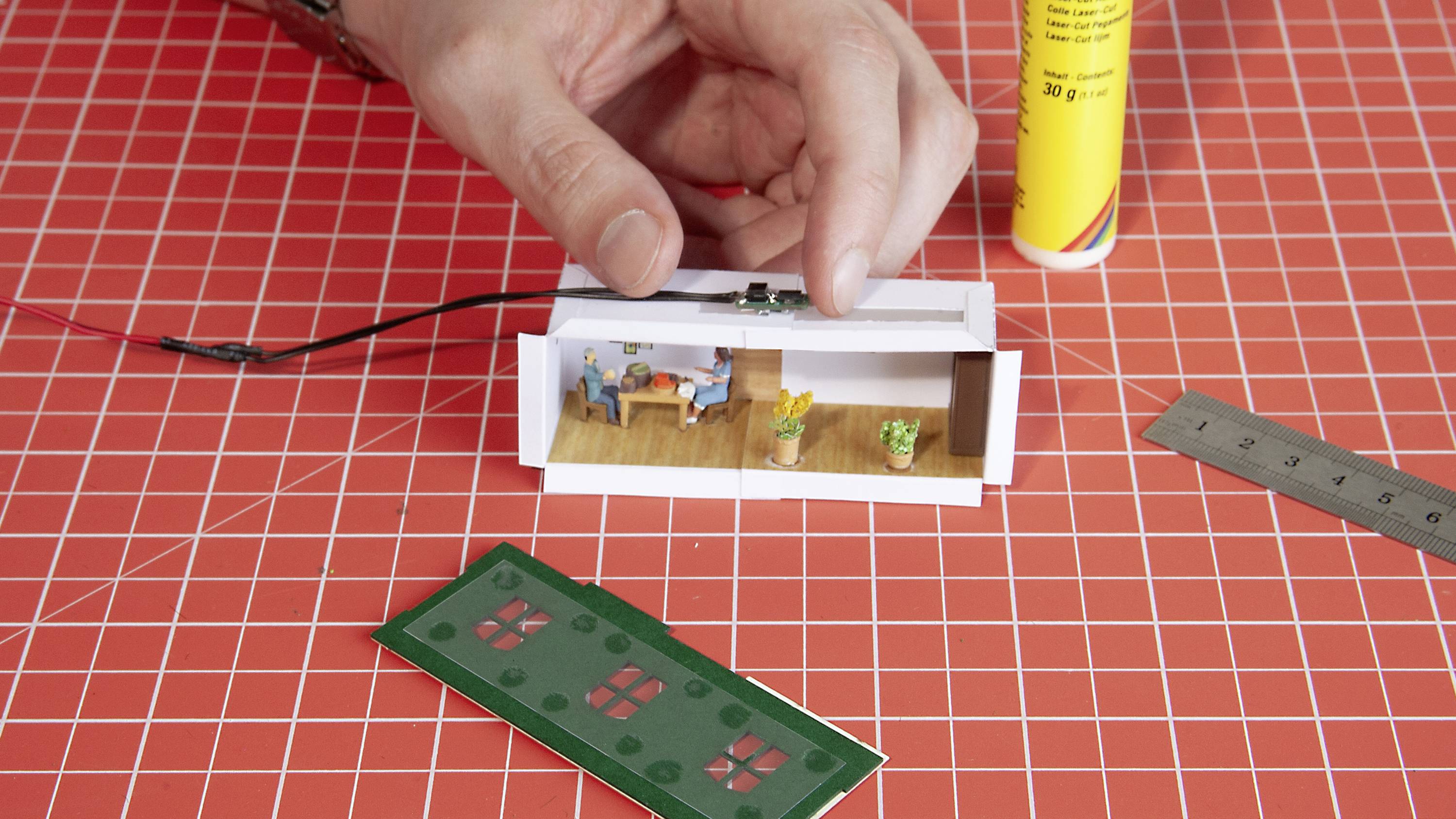 A hand positions miniature interior furnishings in a small model room on a red grid background, with glue and a ruler beside it.