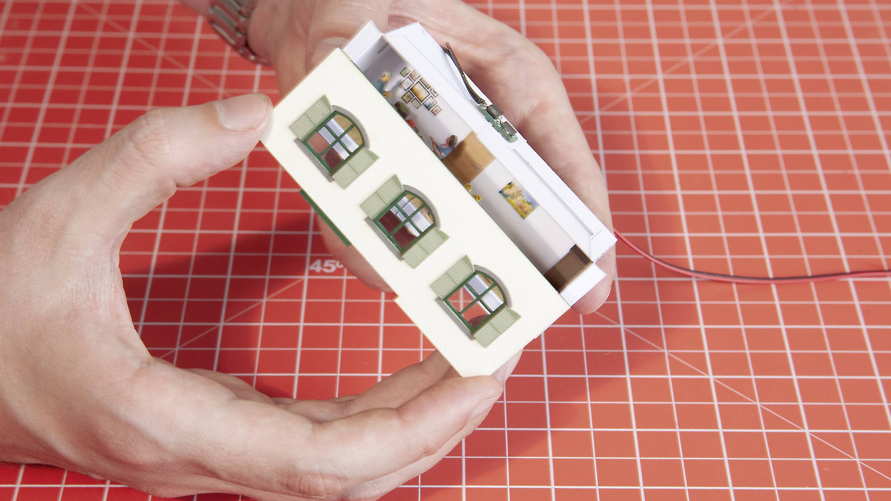 A person is holding a small model house with green windows over a checked background.