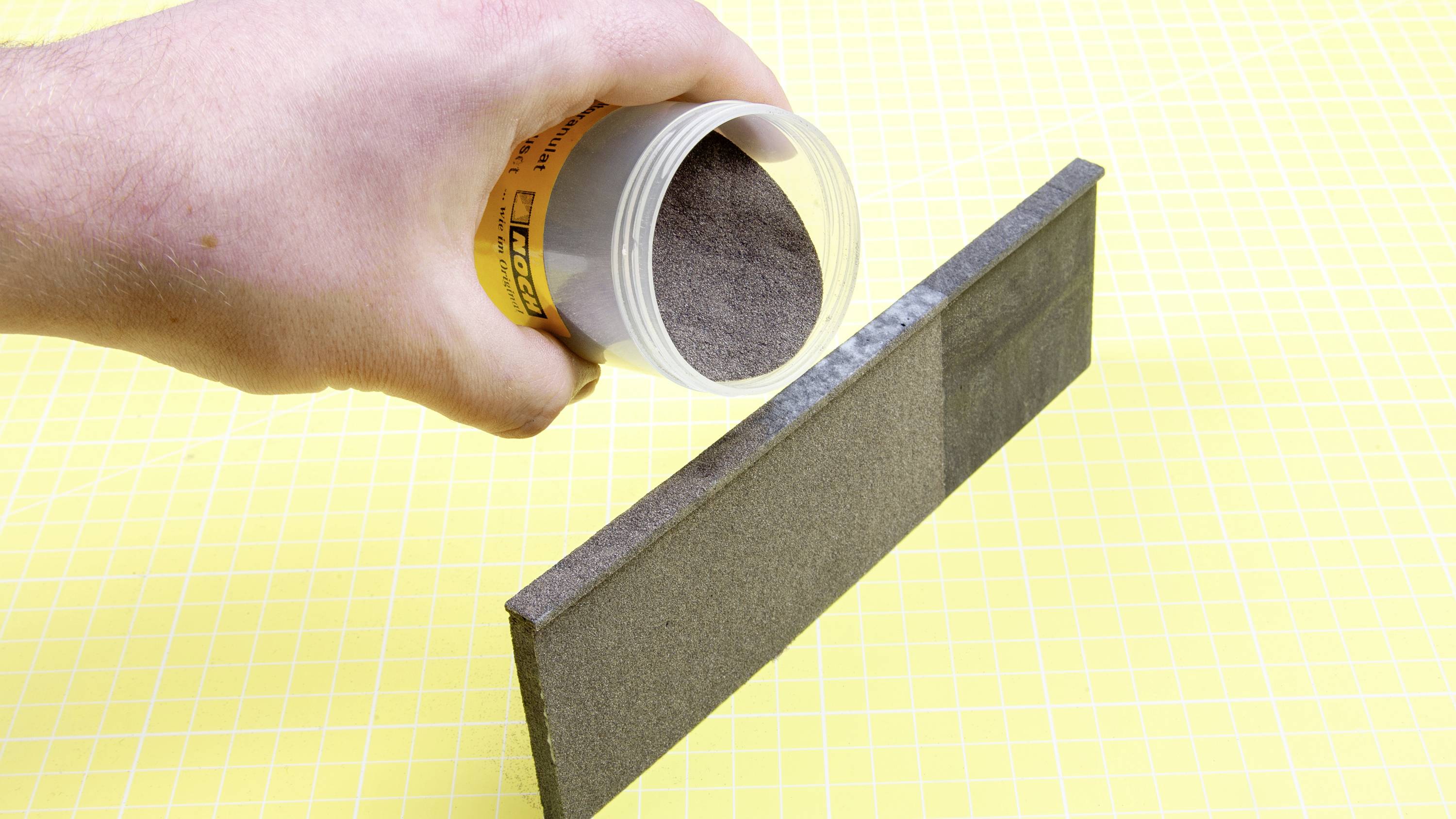 A hand pours grey powder from a container onto a thin, rectangular surface standing vertically. Yellow background.
