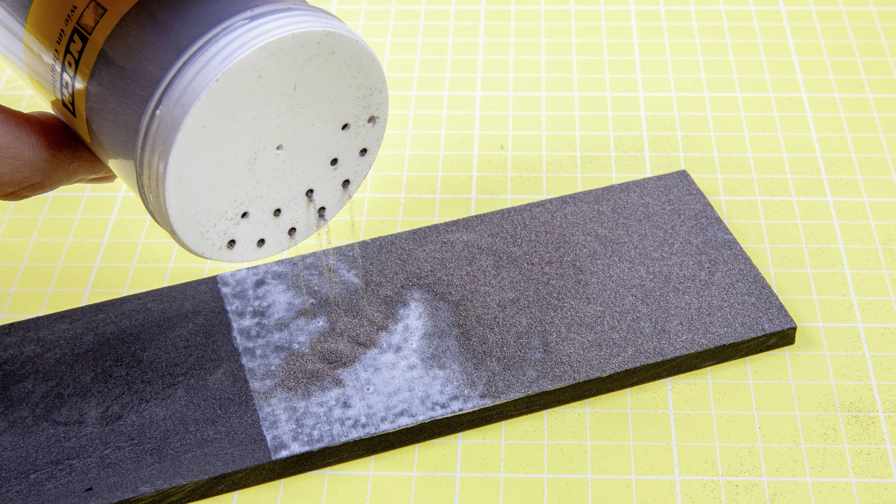 'A hand sprinkles powder onto a sanding block against a yellow, checked background. One half of the block shows powder, the other half does not.'