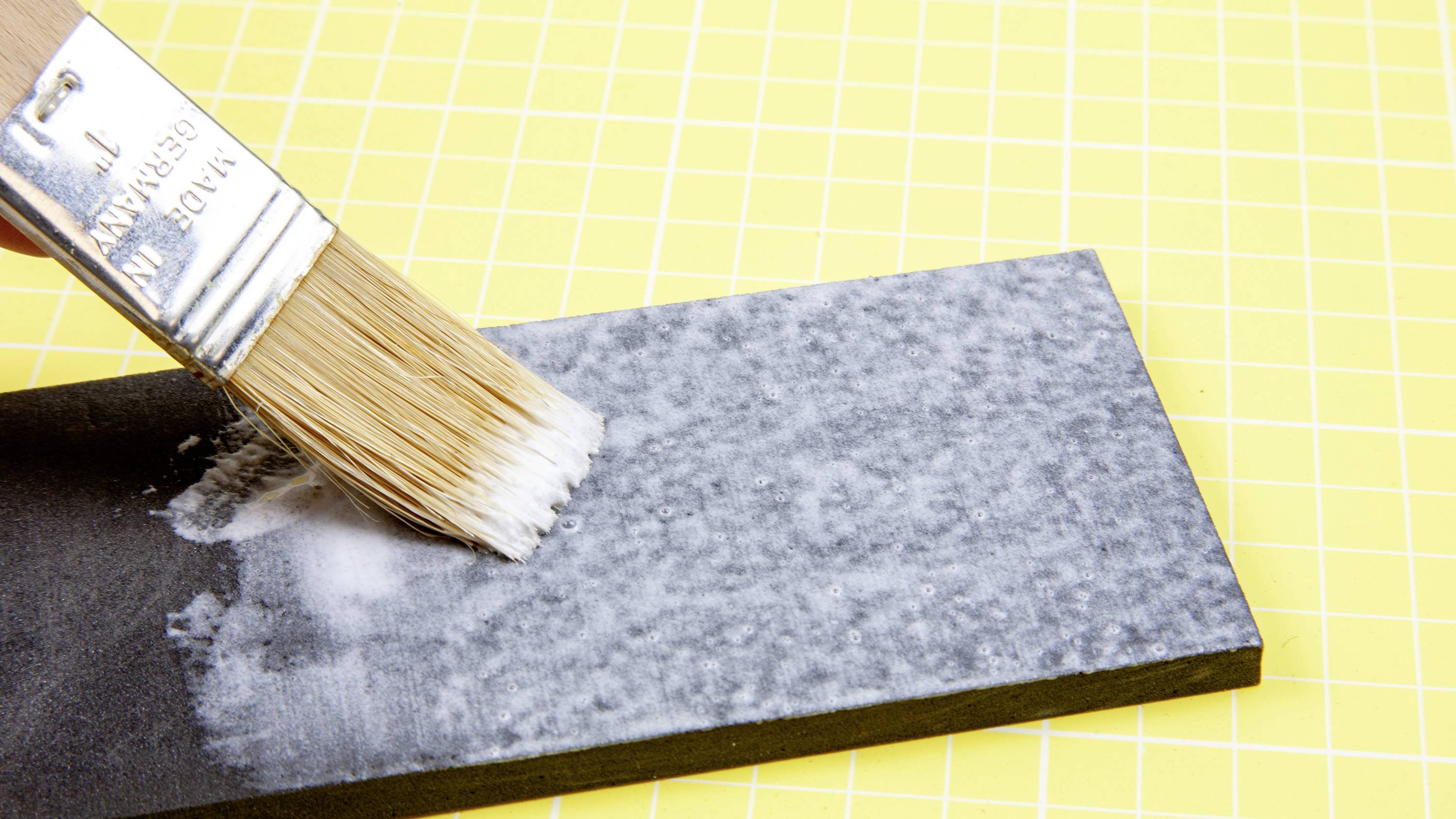 A paintbrush applies white paint onto a black rectangle on a yellow background with a grid.
