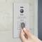 A hand holds a key fob up to an access control system on a wall. The system has a camera and an RFID reader.