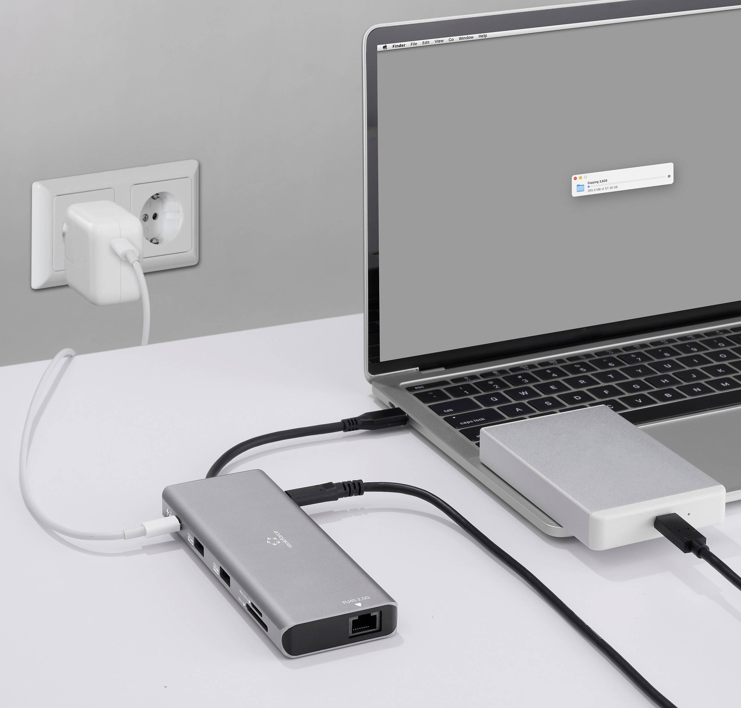A laptop is connected to an external hard drive and a power socket via a USB-C hub and multiple cables. A pop-up is visible on the display.