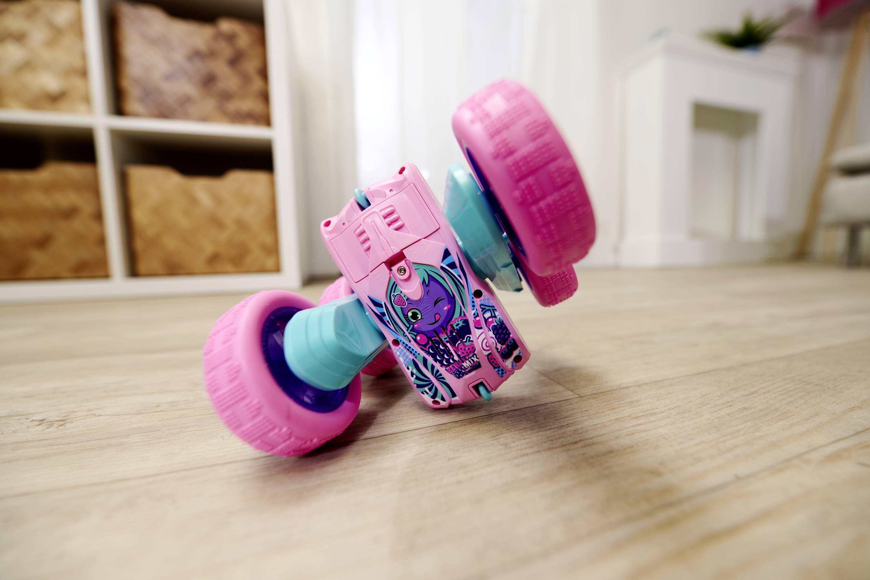 A colourful toy vehicle with large pink wheels lies on its side on a wooden floor in a bright room.