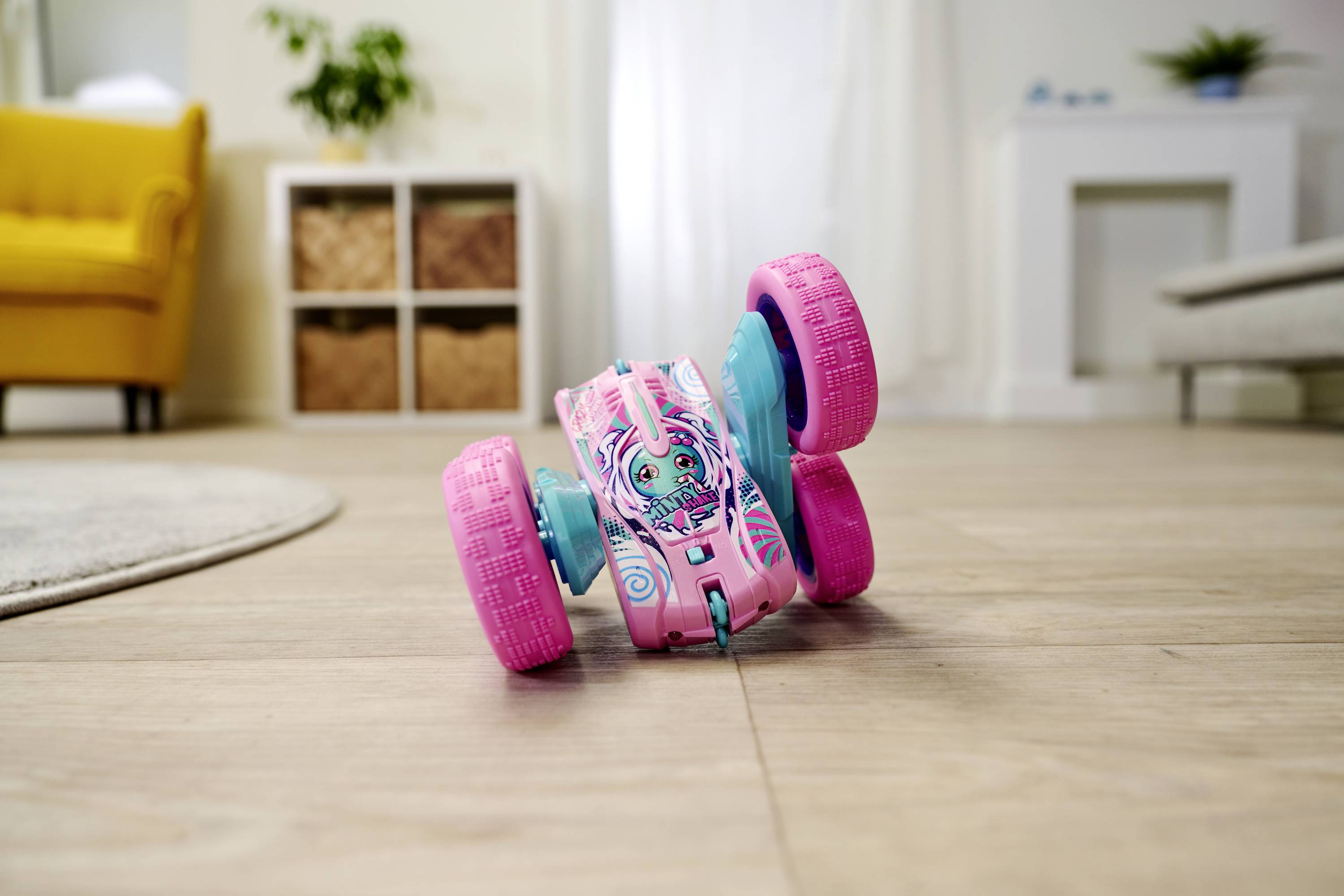 A remote-controlled toy car with pink and blue wheels is standing on a wooden floor in a room with a yellow armchair in the background.