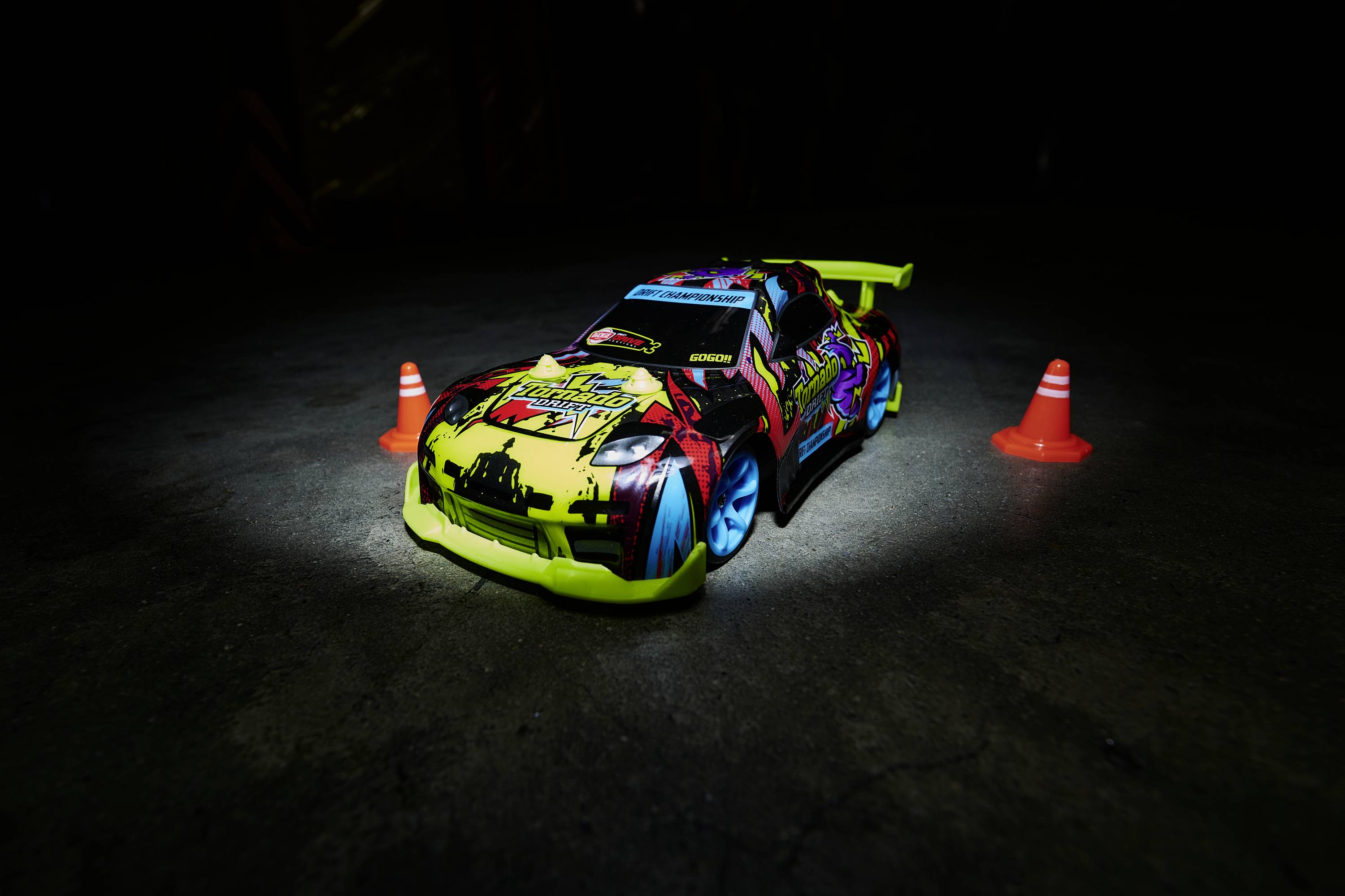 A brightly painted toy car drives across a smooth surface between two small orange traffic cones in dim light.