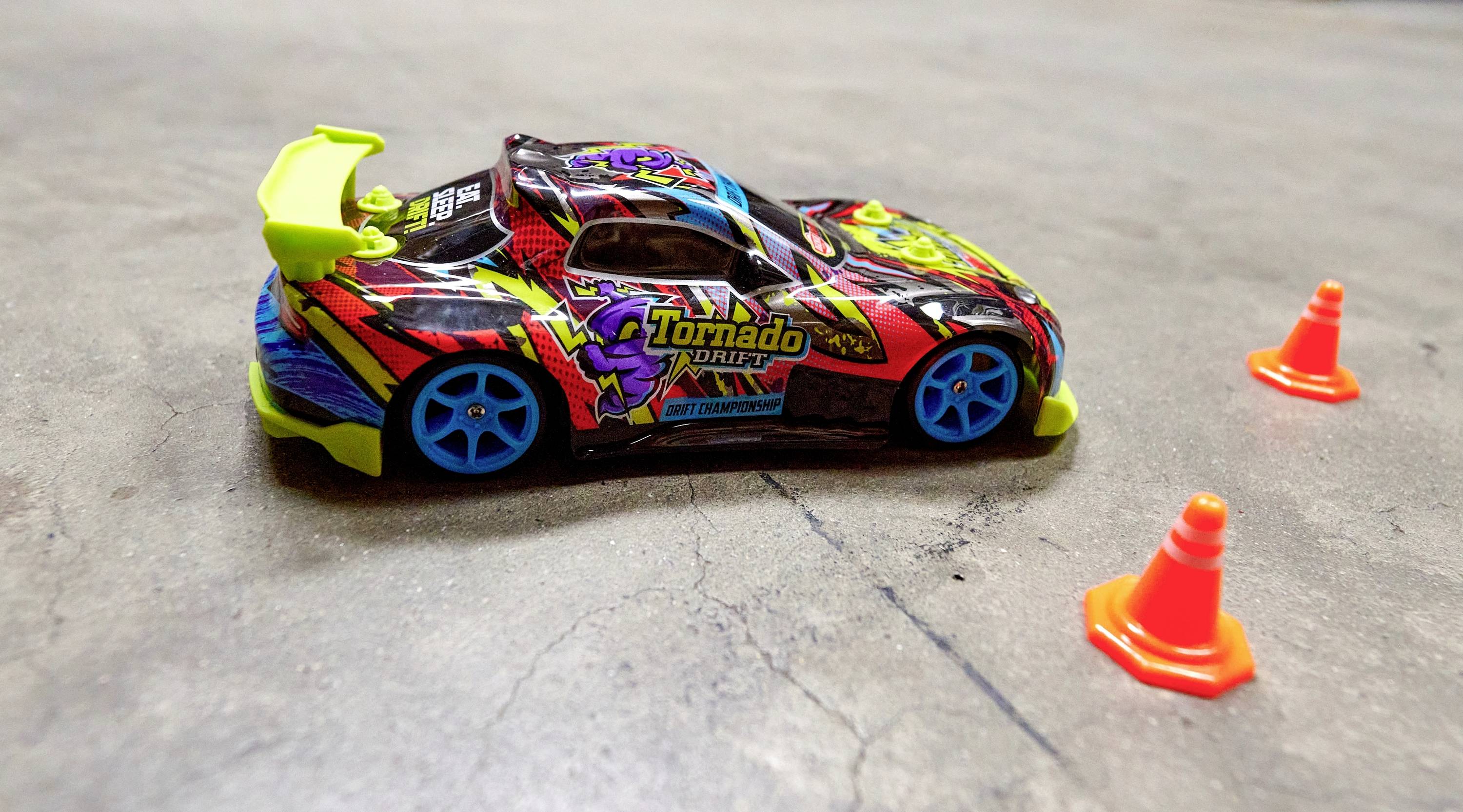 A toy car with a colourful design and the inscription 'Tornado Drift' is standing on a floor, flanked by two small traffic cones.