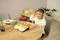 A child sits at a table wearing headphones. Children's books and wooden toys are laid out in front of them. Plants are visible in the background.