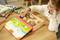 A child is playing with a wooden puzzle at the table. Numerous wooden pieces fit into a colourful board. Books and headphones are in the background.