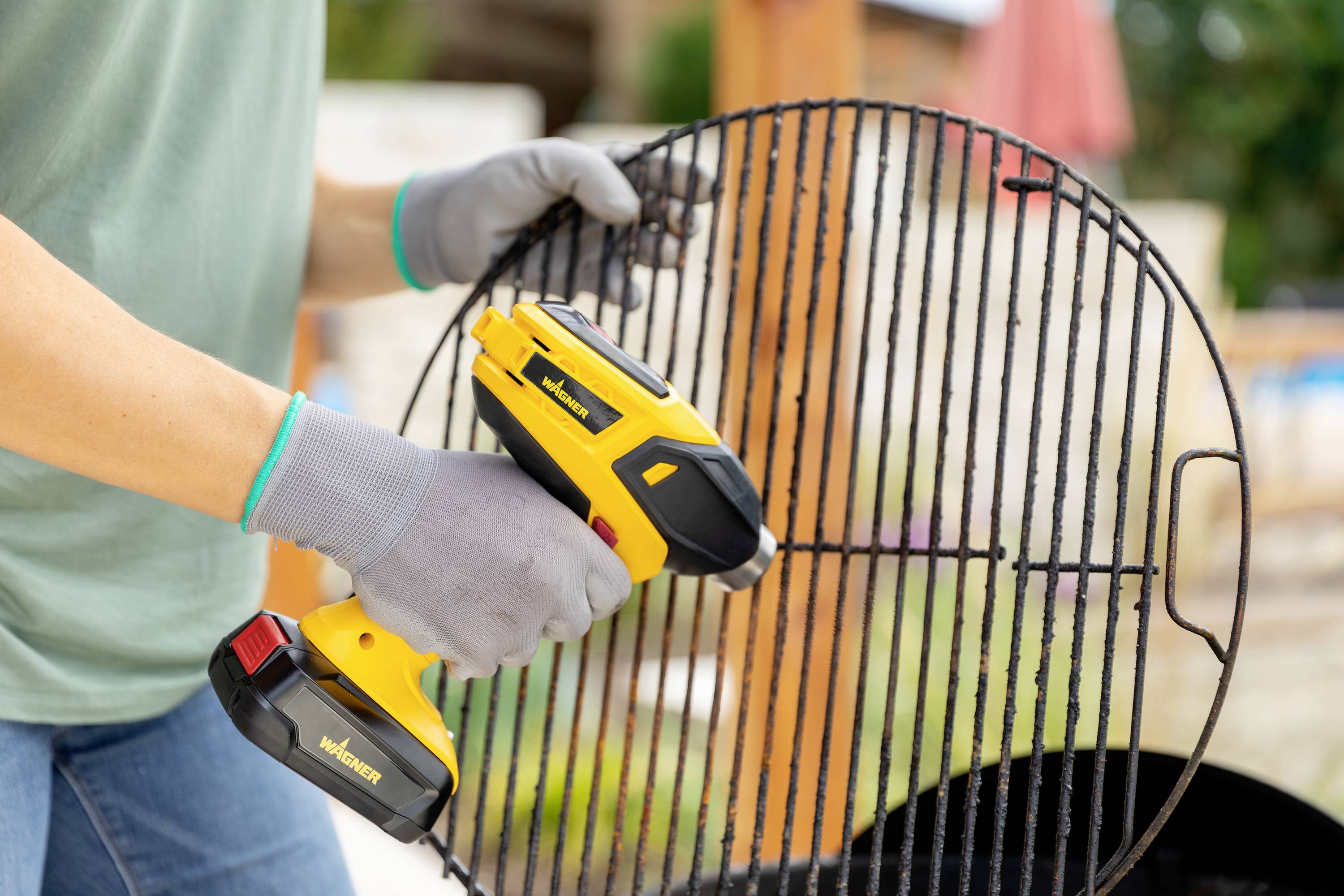 A person wearing gloves holds a yellow hot air dryer and cleans a barbecue grill. A barbecue is visible in the background.