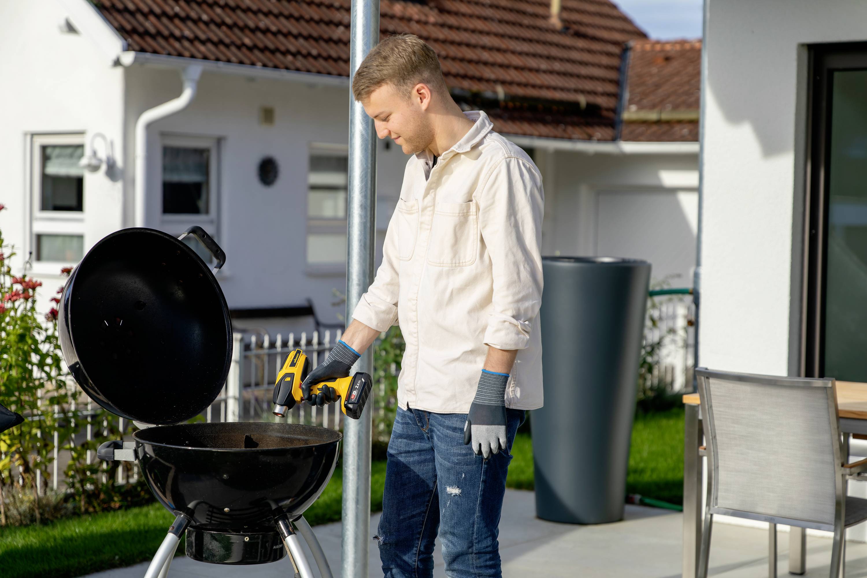 A man is standing outside grilling and handling tools. Houses and a garden are visible in the background.