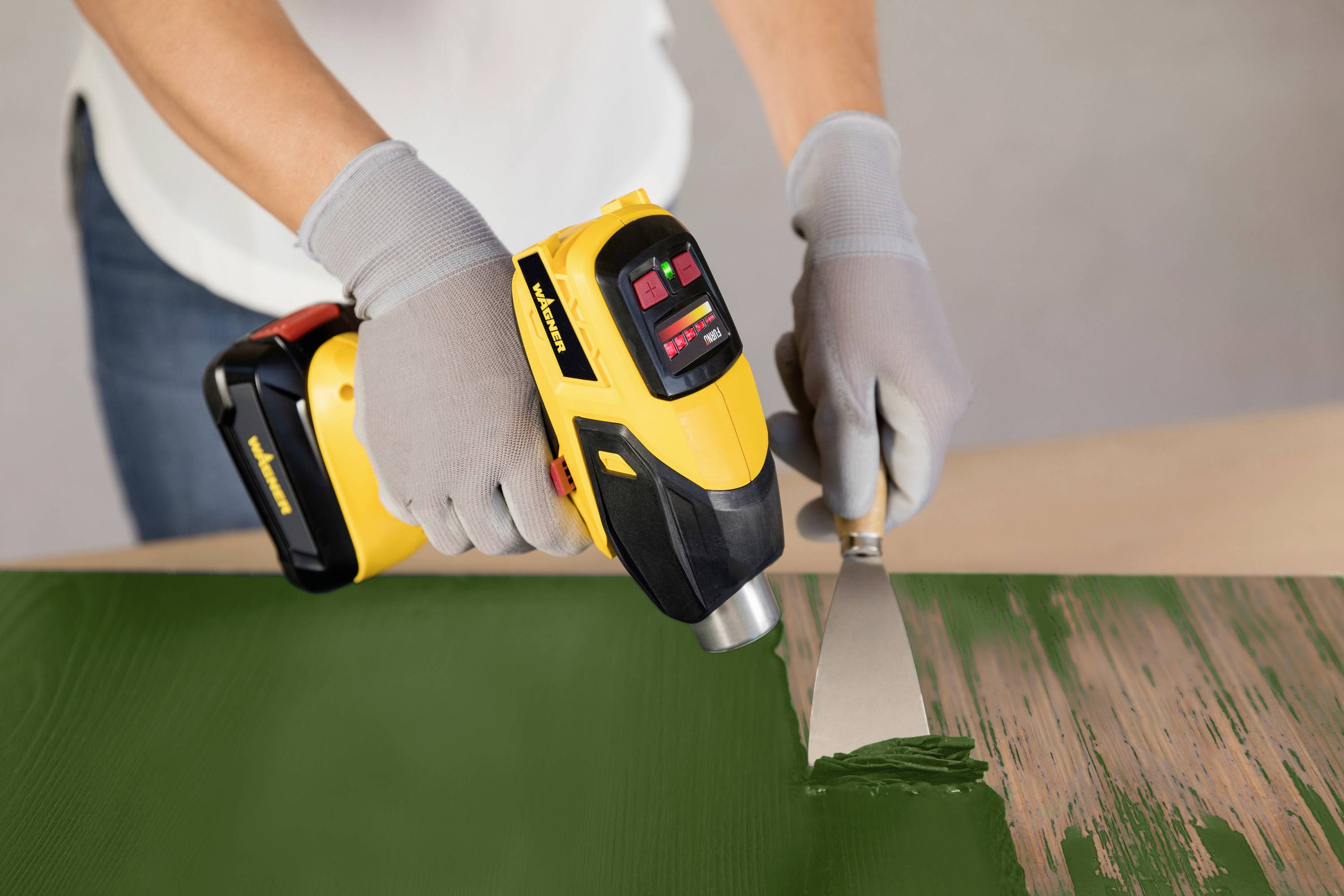 A person is removing old paint from a wooden surface using a hot air gun and a scraper.