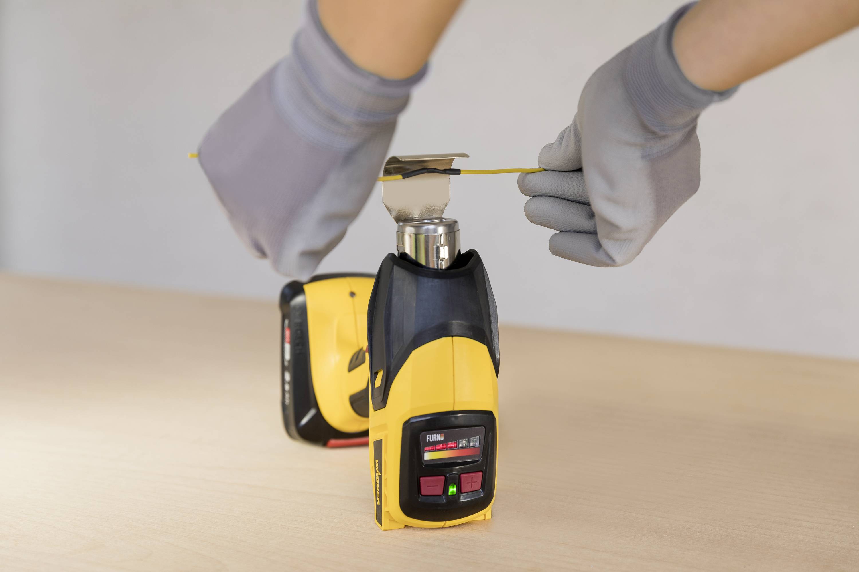 A person wearing grey gloves is stretching a yellow measuring tape over a yellow digital device with a red display on a table.
