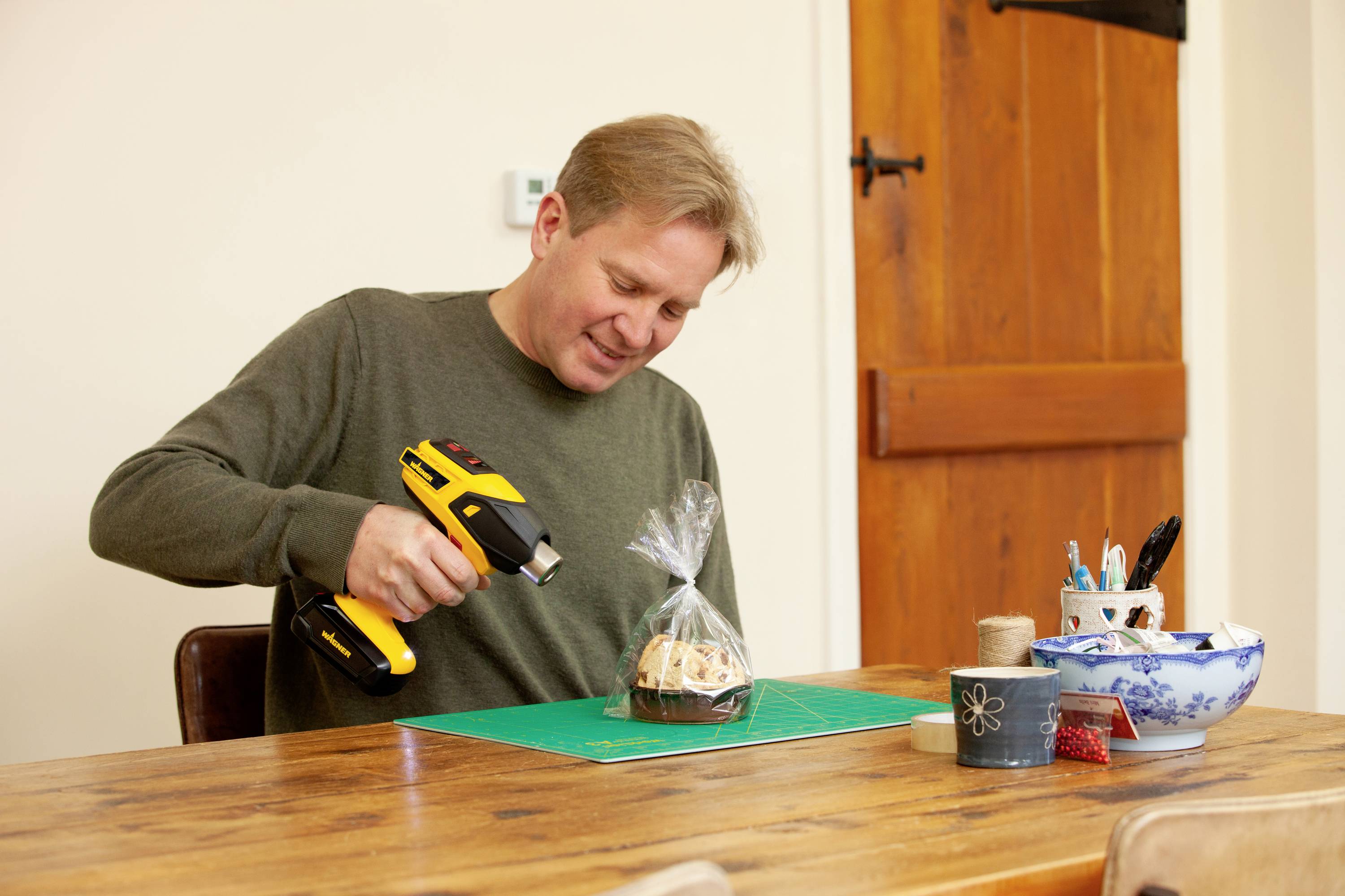 A person is using a heat gun to shrink plastic film around a small package on a table with craft materials.