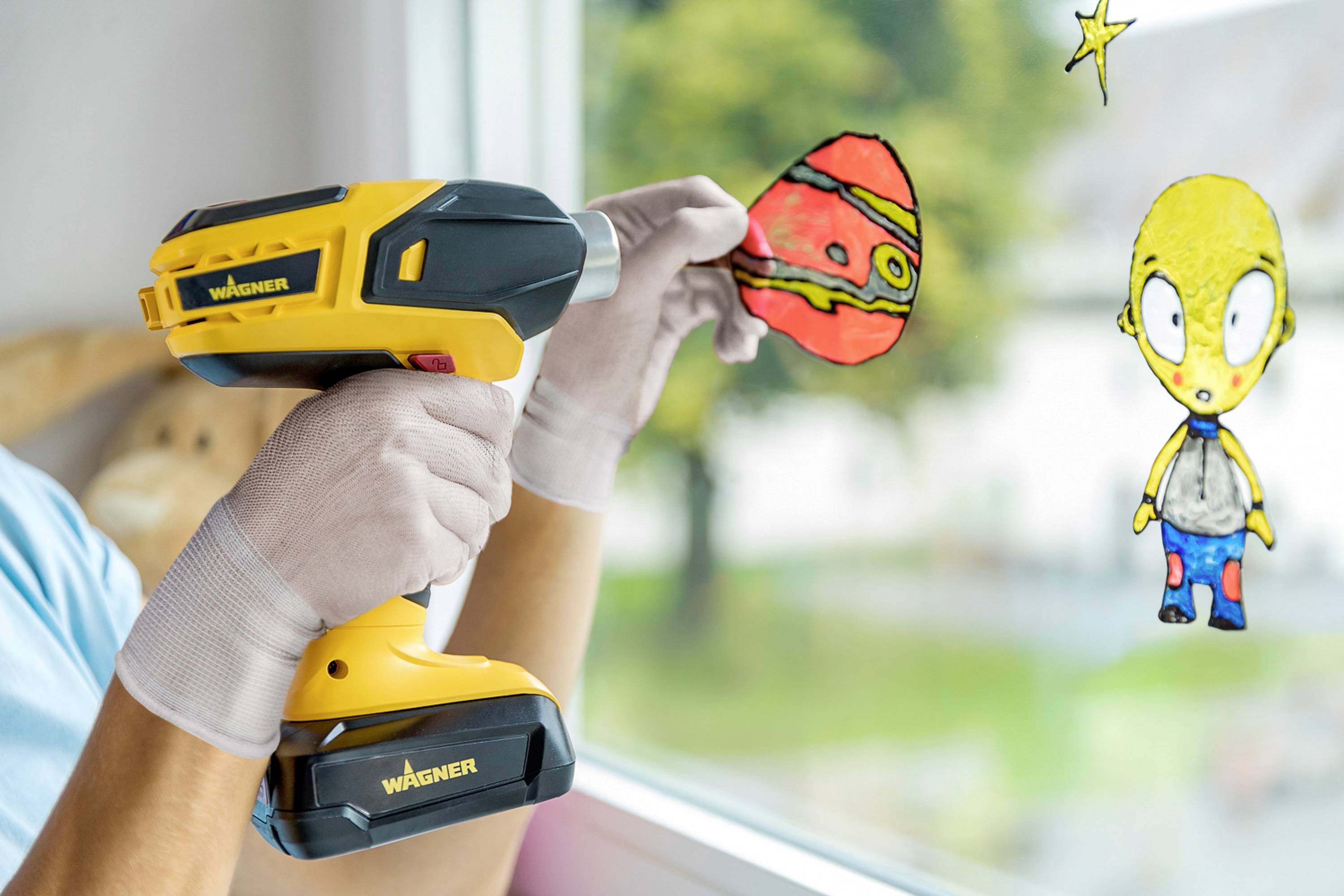A person is holding a hot glue gun and attaching a colourful paper picture to a window. A green landscape is visible in the background.