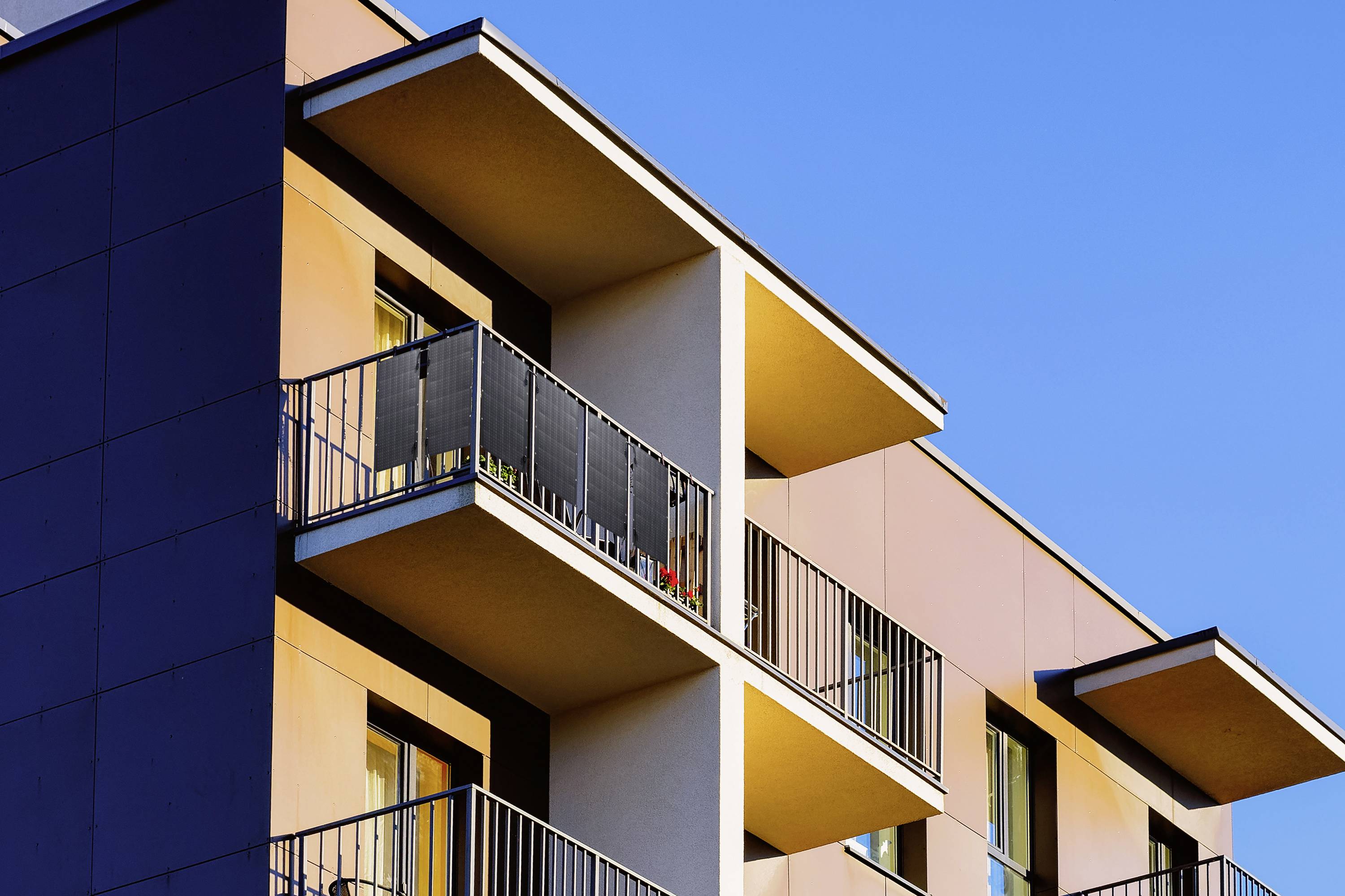 A modern residential building with multiple balconies, illuminated by the evening sun. The sky is clear and blue.