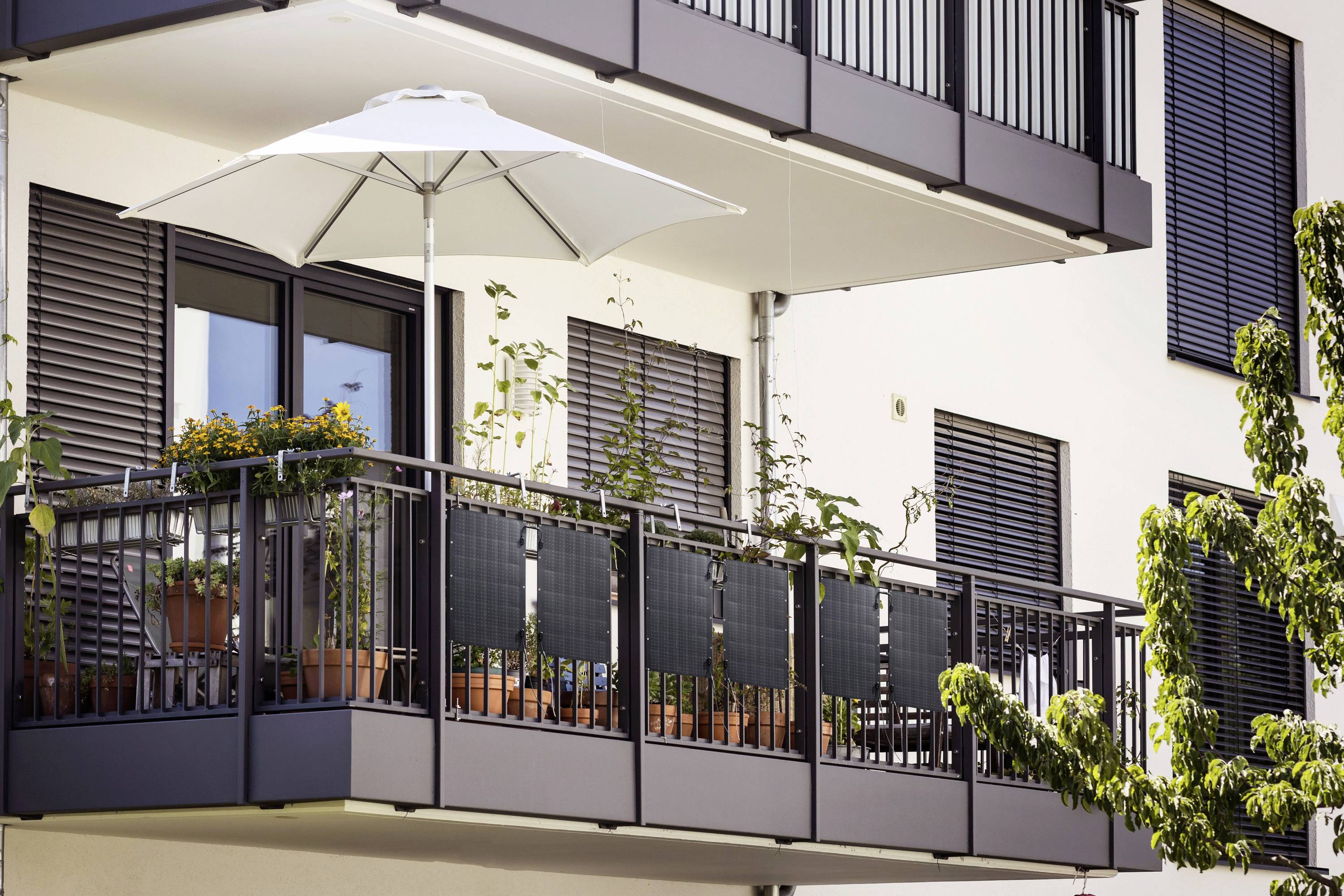 Balcony of a modern apartment with potted plants and an open parasol, surrounded by trees.