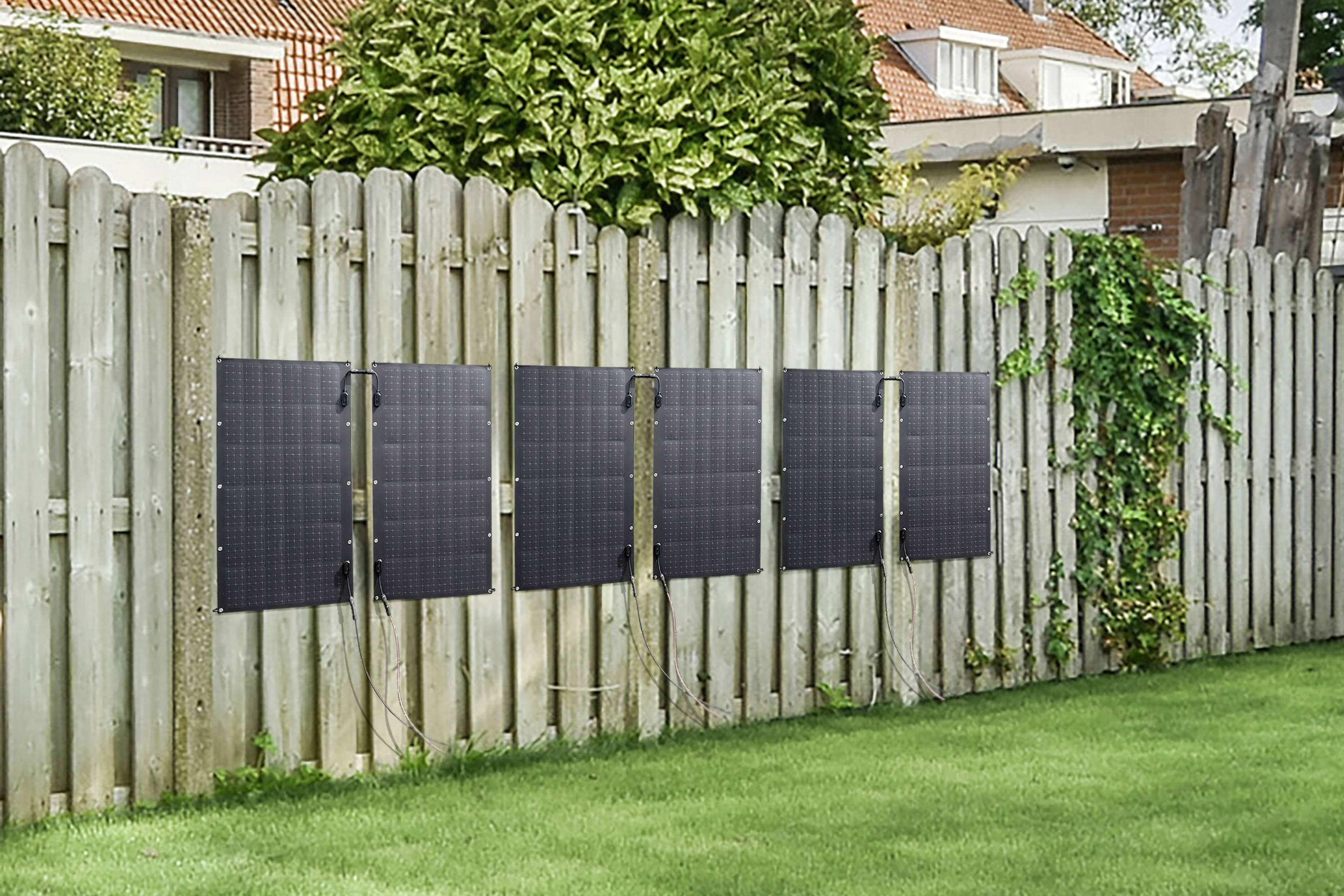 Five solar modules are mounted on a wooden fence in the garden. Houses and trees can be seen in the background.