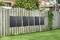 Five solar modules are mounted on a wooden fence in the garden. Houses and trees can be seen in the background.