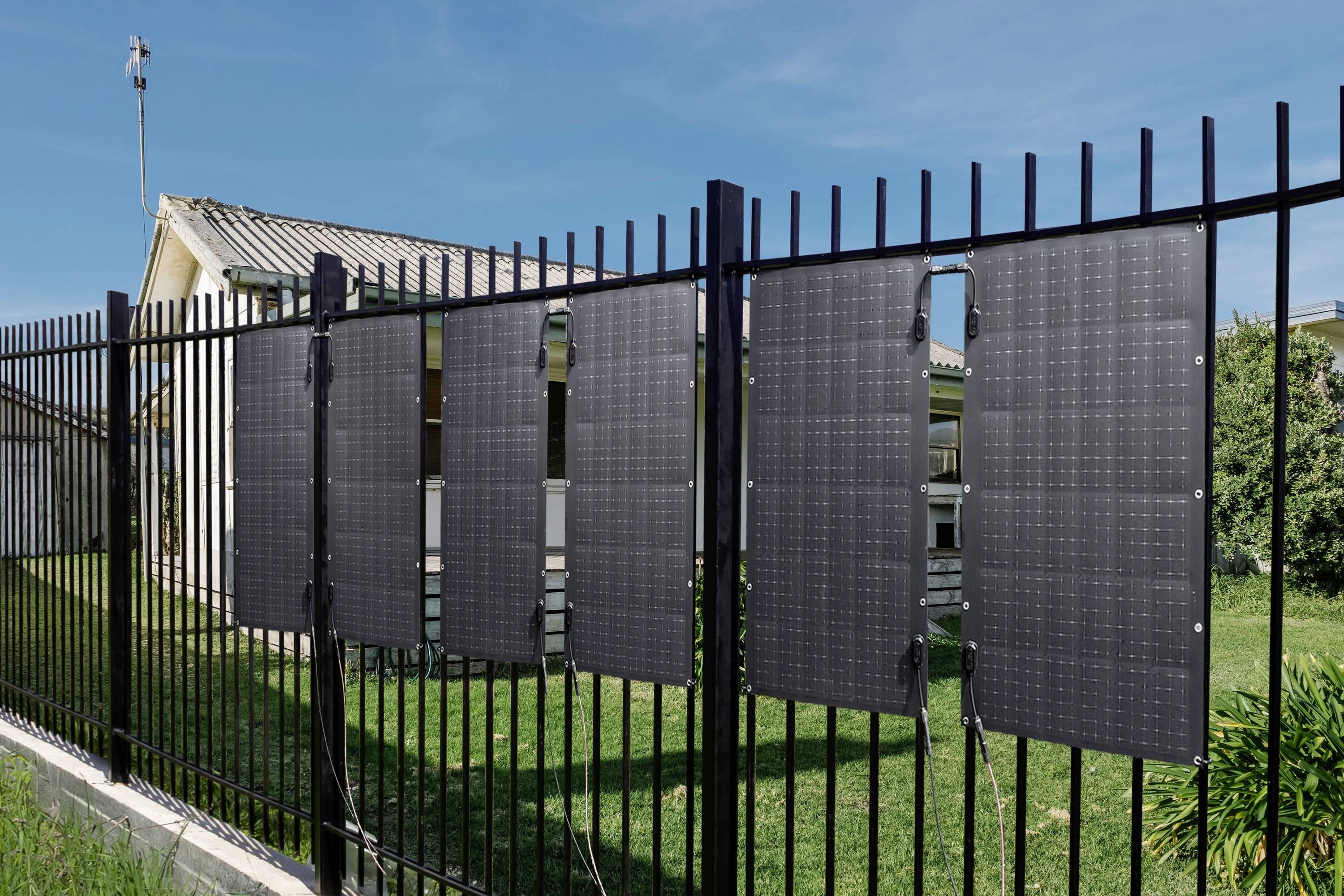 Solar panels are attached to a metal fence, with a house in the background. The scene depicts sustainable energy use in a residential area.