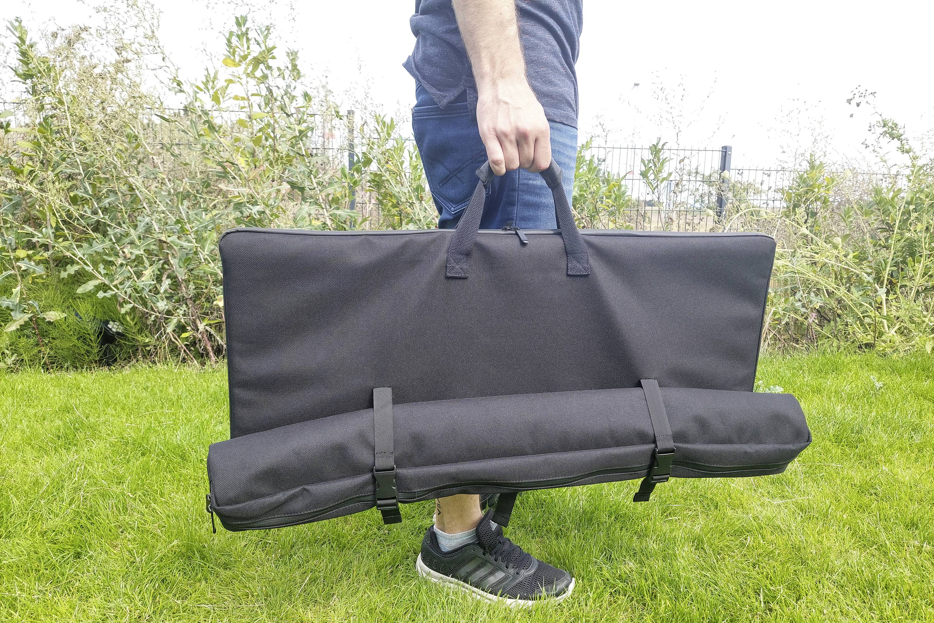 A person is carrying a rolled-up black bag and a large, rectangular bag over a green lawn.