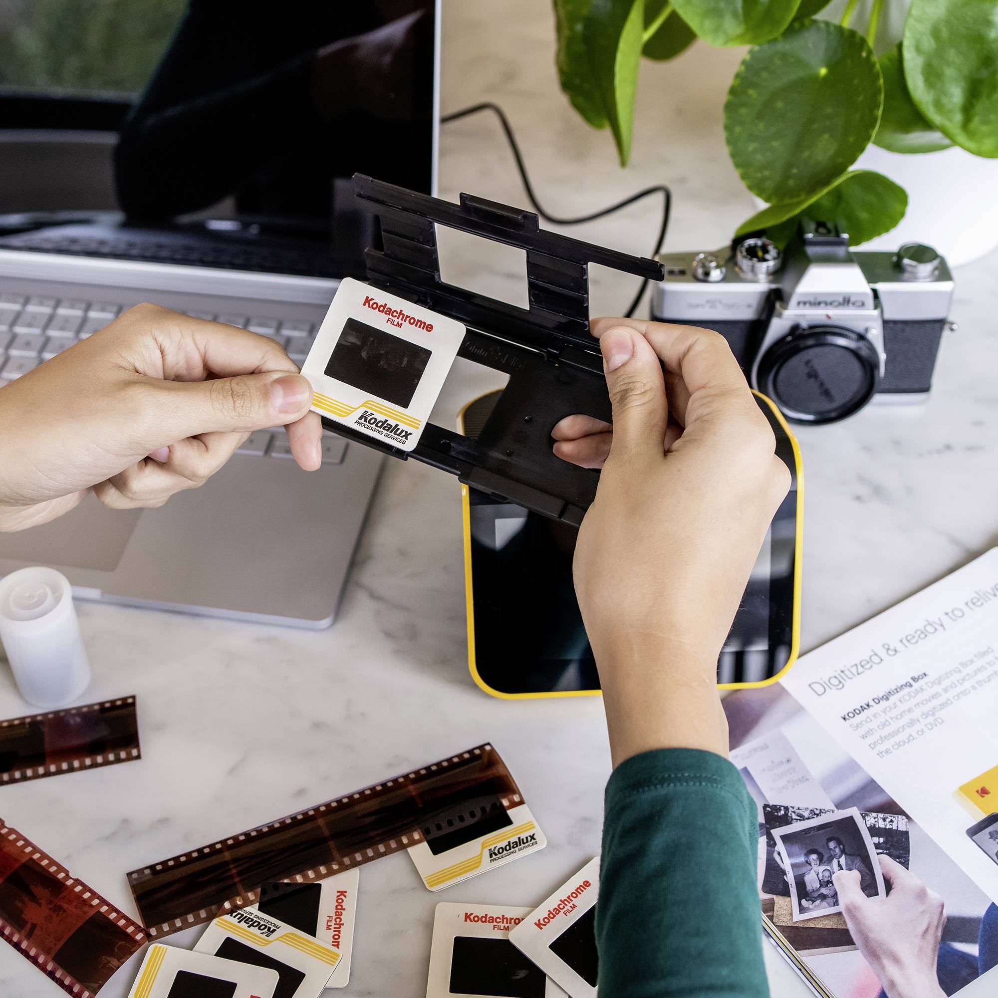 A person is holding a slide frame with a Kodachrome slide. More slides and an old camera are lying on the table. A laptop can be seen in the background.