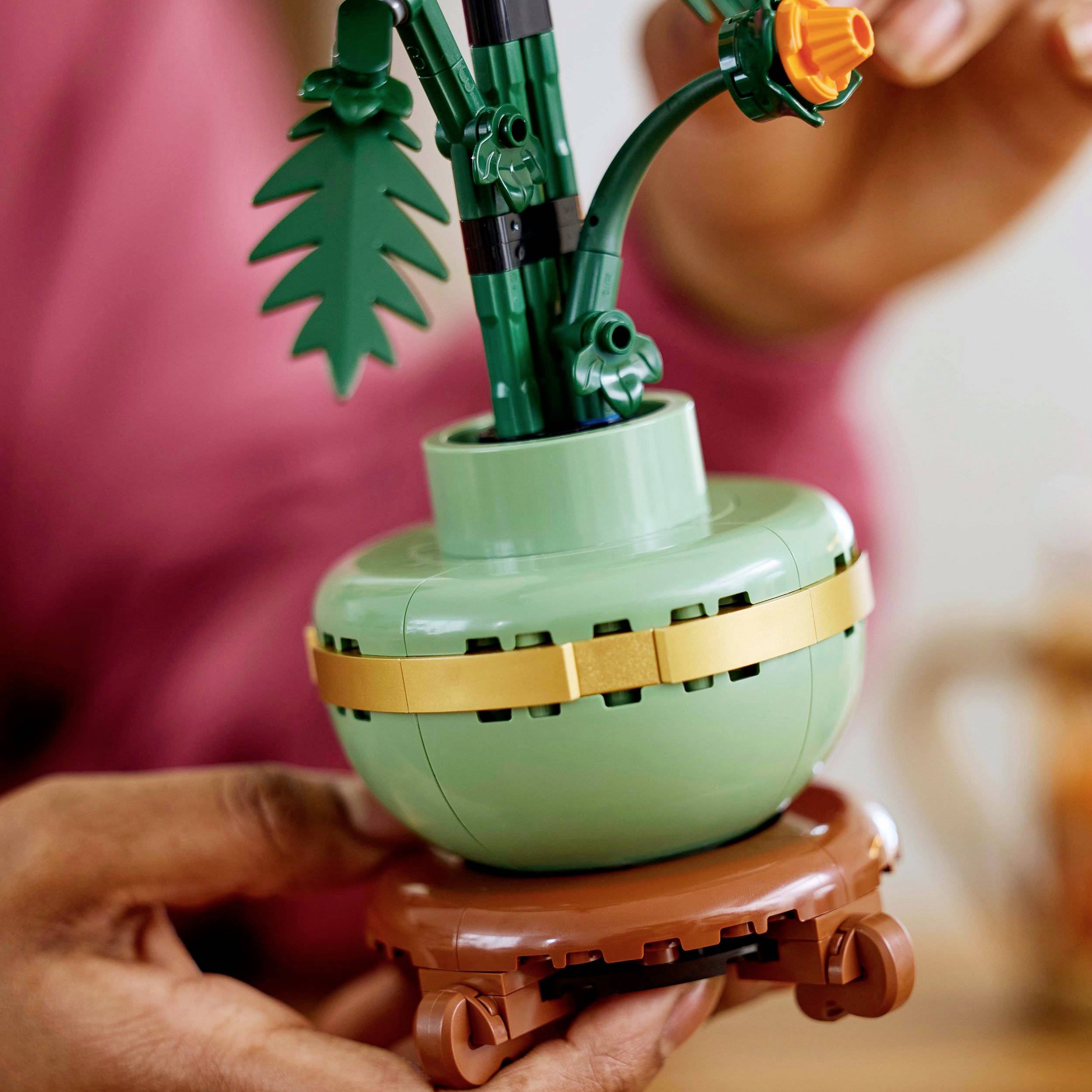 A person is holding a Lego model of a plant with green leaves and an orange flower in a green pot on a brown stand.