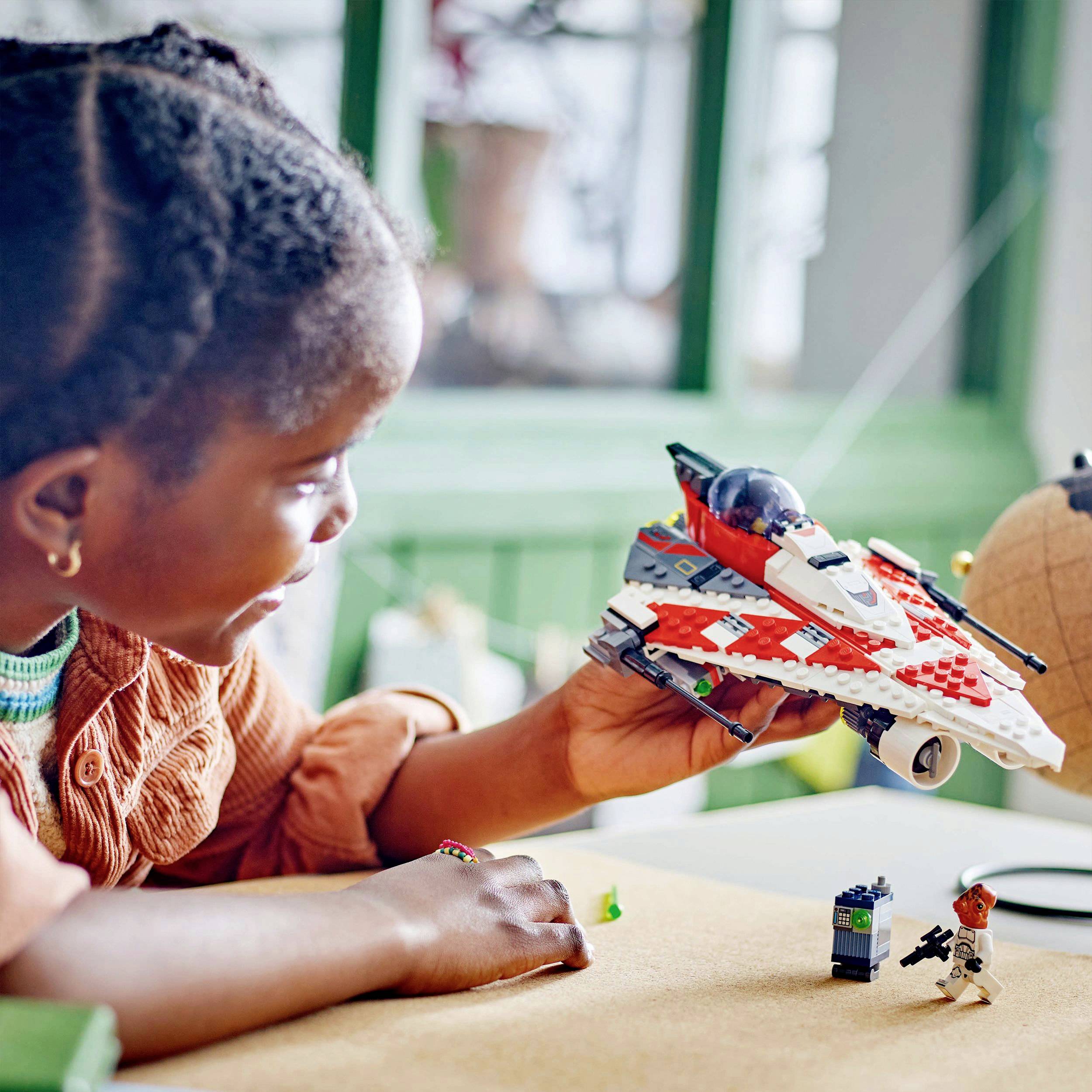 A child is playing with a homemade toy spaceship made from Lego bricks. A small Lego figurine stands beside it on the table.