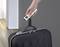 A hand is holding a digital luggage scale attached to a black suitcase handle. The background is a modern interior.