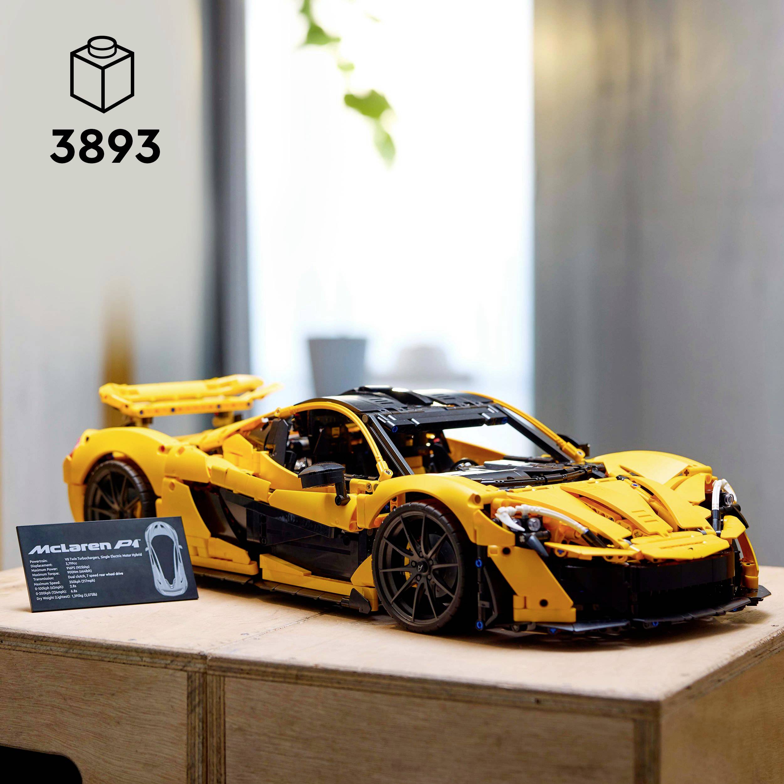 A yellow model car at scale on a wooden table, with an information label beside it. In the top left is the symbol of a building block with the number '3893'.