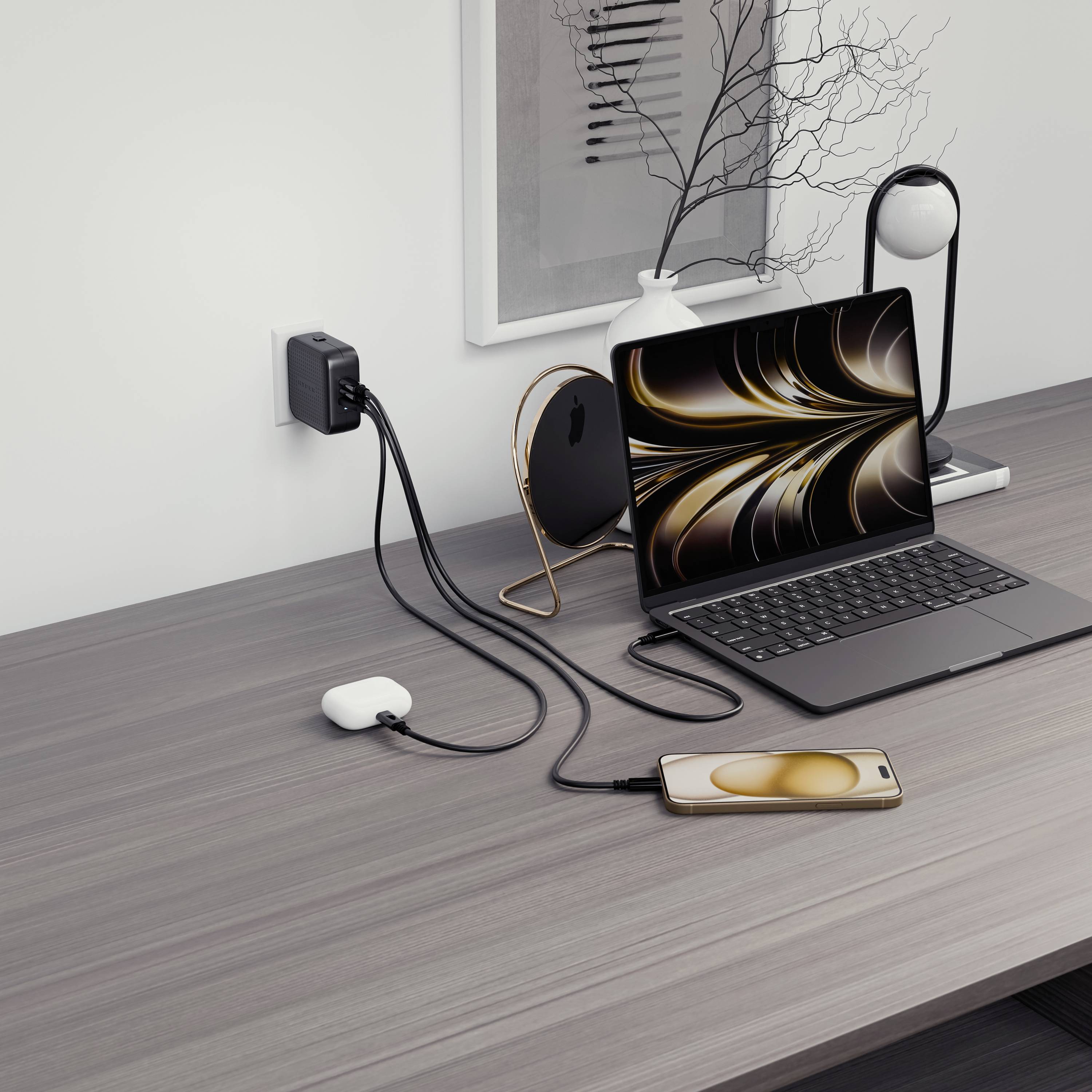 A desk with a laptop, a mobile phone and charging cables; the laptop is connected to the mobile phone; modern charger plugged into the socket.