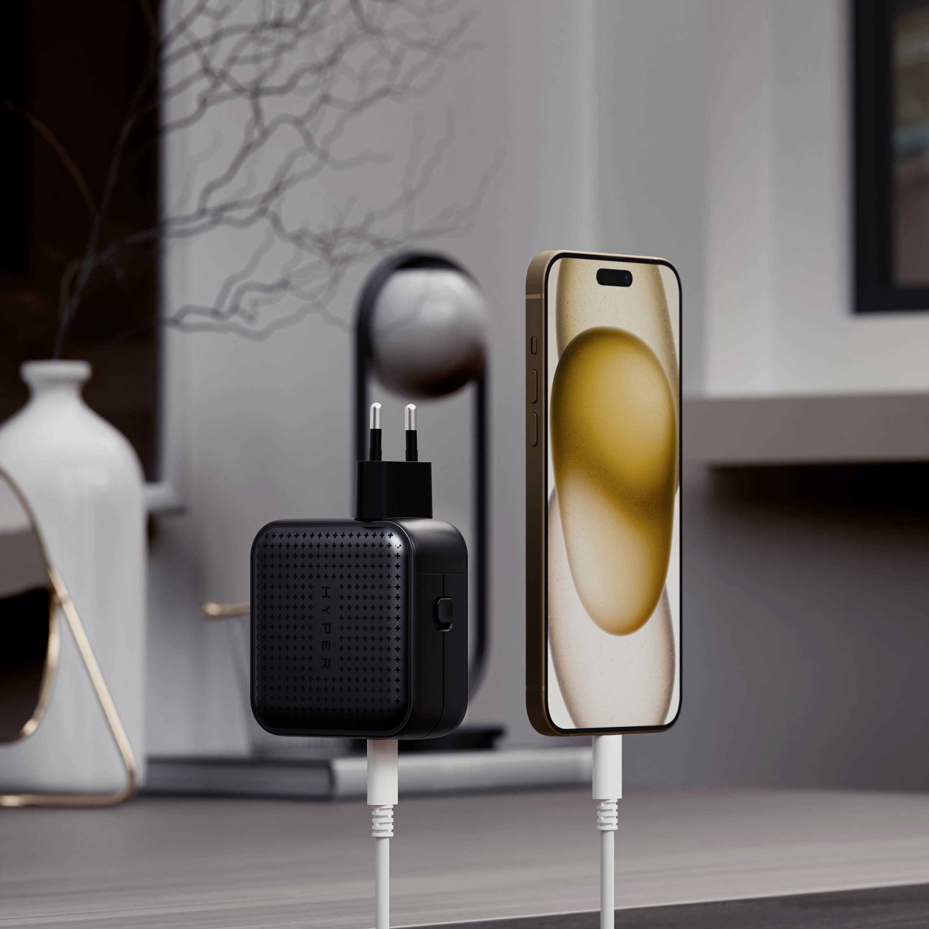 A smartphone is being charged wirelessly. A charger is standing next to it, plugged into a power socket. Modern decor is visible in the background.