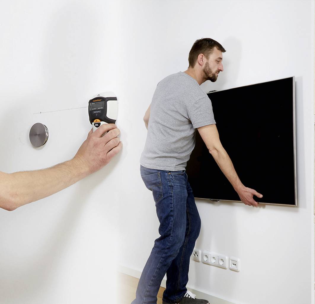 A man is mounting a flat-screen television on the wall. A measuring tape is attached to the wall in the background.