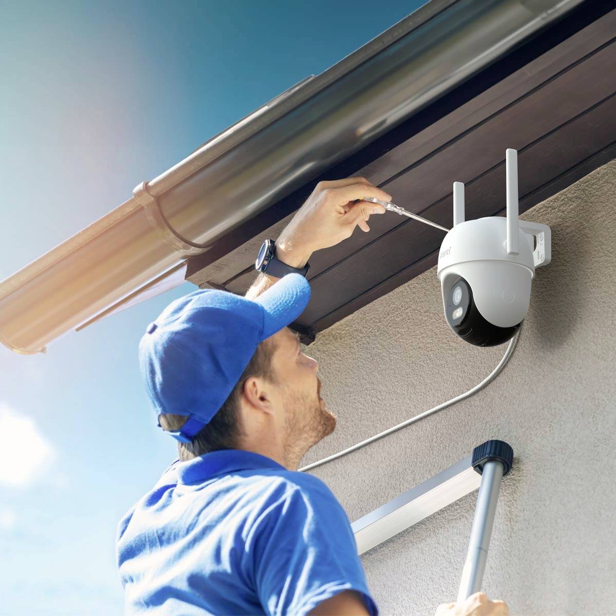 A man in blue workwear is installing a surveillance camera on the house wall while tightening a screw.