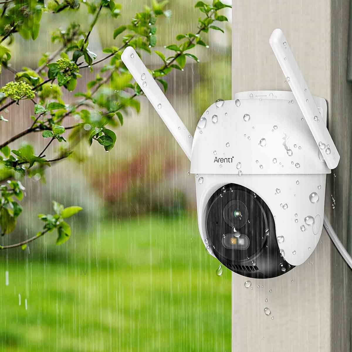 Security camera on a house wall in the rain, with water droplets on the lens, surrounded by green plants.