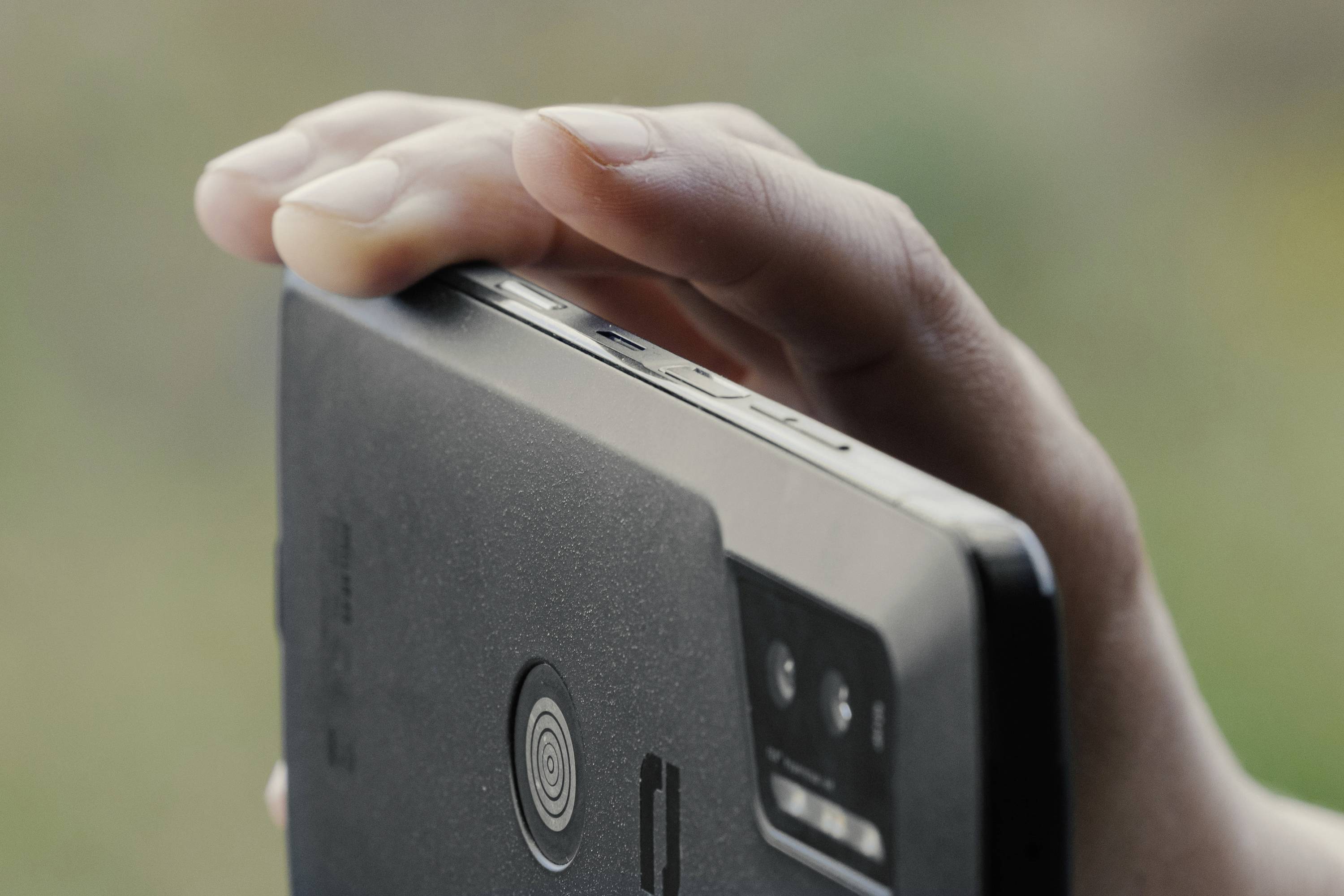 A hand holding a smartphone, viewed from the side. The camera and volume buttons are visible. Background blurred.