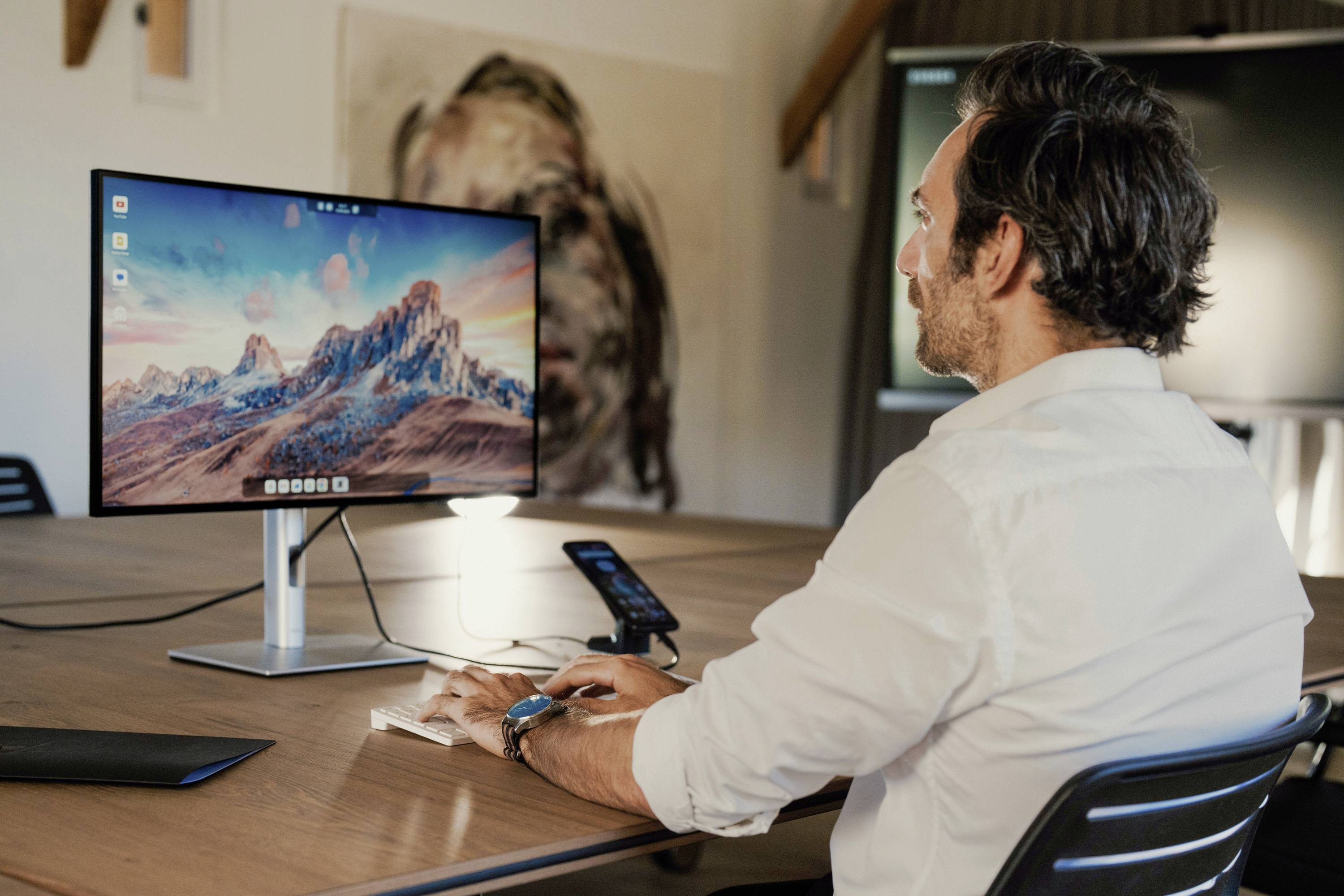 A man is sitting at a desk and working on a computer. A landscape with mountains is displayed on the screen.