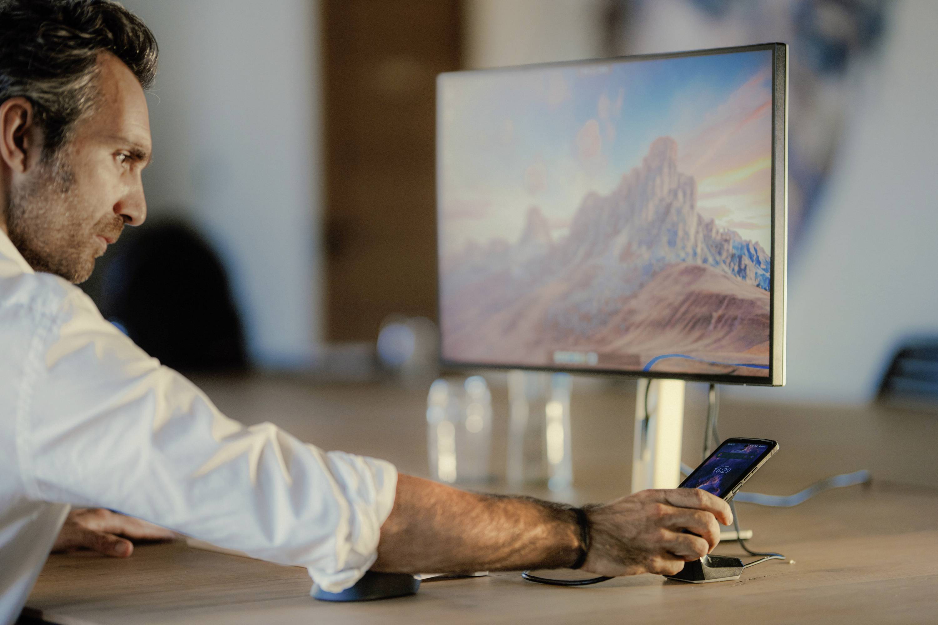 A man is operating a smartphone that is sitting on a table while looking at a large monitor displaying a mountain landscape.