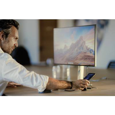 A man is operating a smartphone that is sitting on a table while looking at a large monitor displaying a mountain landscape.