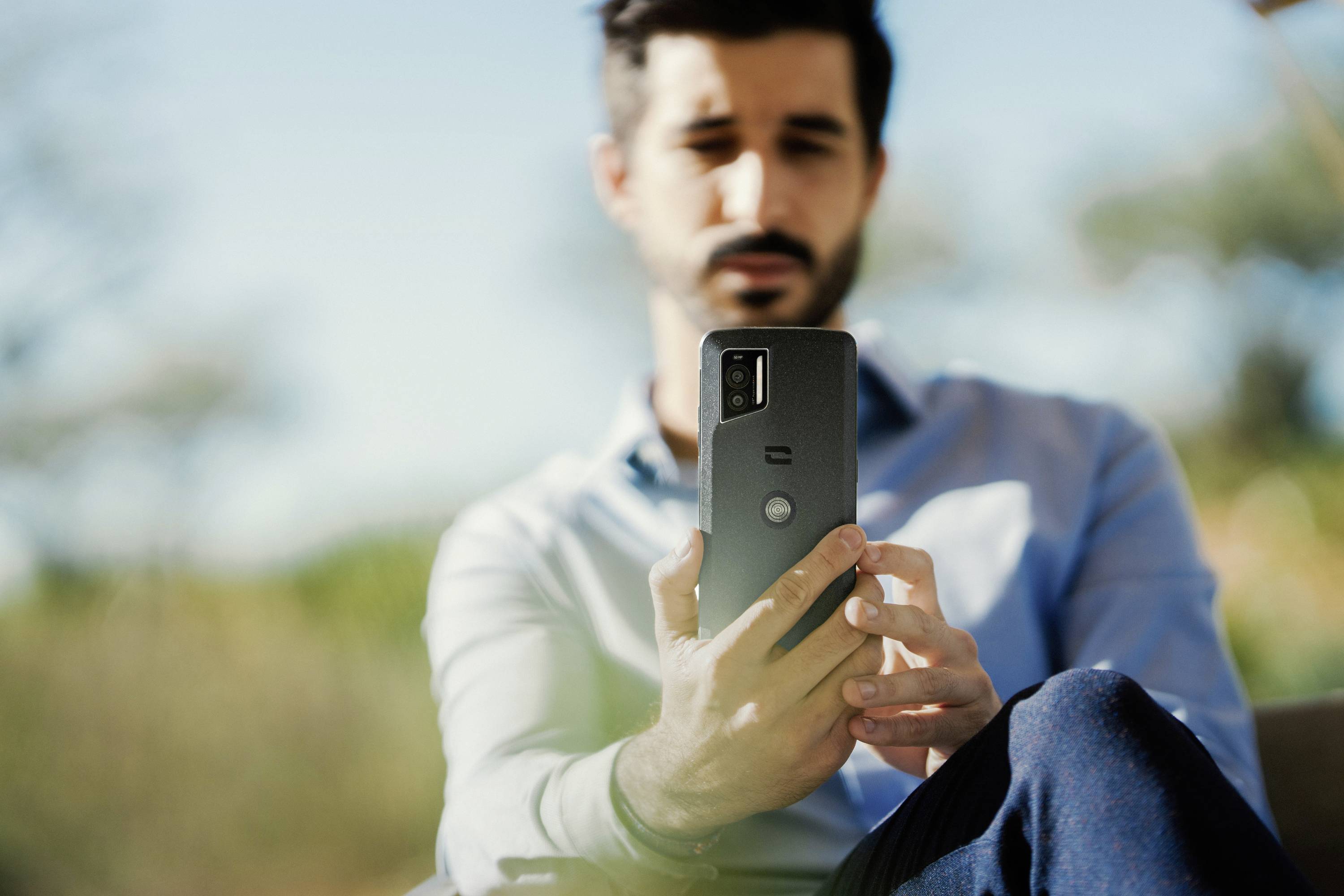 A man outdoors is holding a smartphone in his hand. The background is blurred, showing trees and a blue sky.