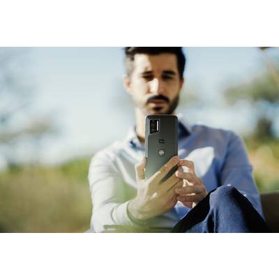 A man outdoors is holding a smartphone in his hand. The background is blurred, showing trees and a blue sky.